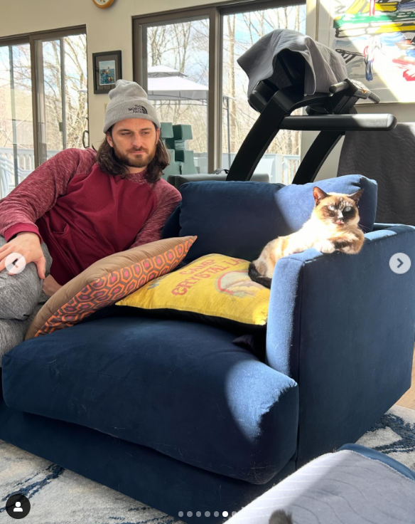 Alex Baglieri sits on a couch with their pet cat basking in the sun, posted in March 2024 | Source: Instagram/em_chases