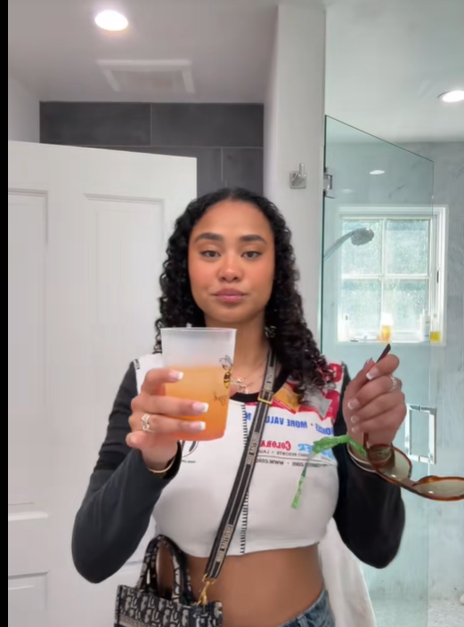 Brianna McKnight stands in a bathroom holding a clear plastic cup with an orange-colored drink in one hand and sunglasses in the other, from a post dated February 8, 2025. | Source: TikTok/brimcknighttt
