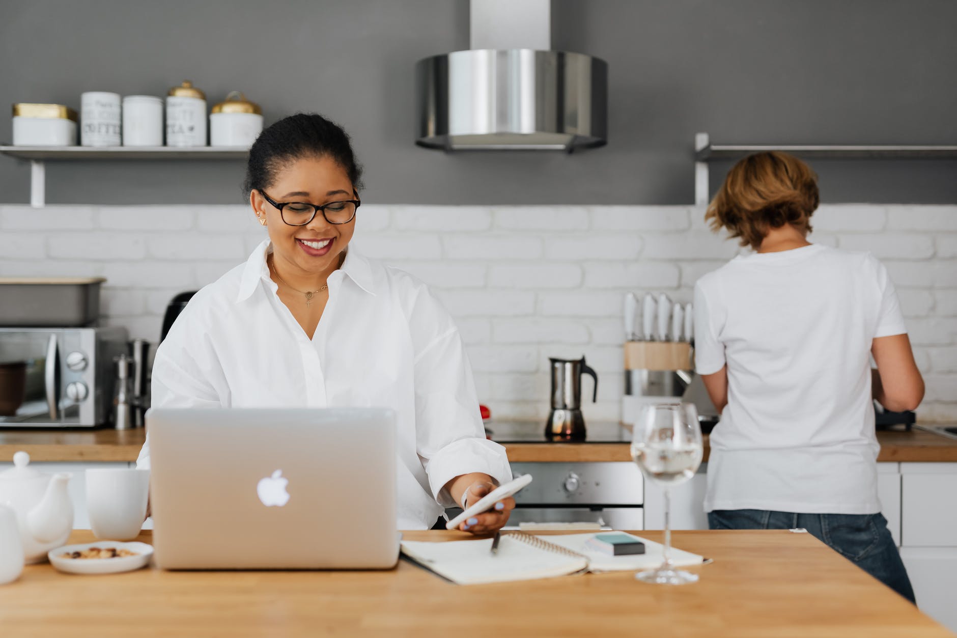 A smiling woman holding a cellphone and working on her laptop on a kitchen counter | Source: Pexels