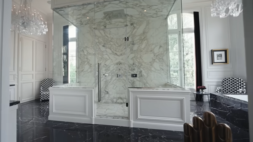 The lavish master bathroom boasts bookmatched white marble walls, a massive glass shower enclosure, black marble floors, and sparkling chandeliers. | Source: YouTube/@Archdigest