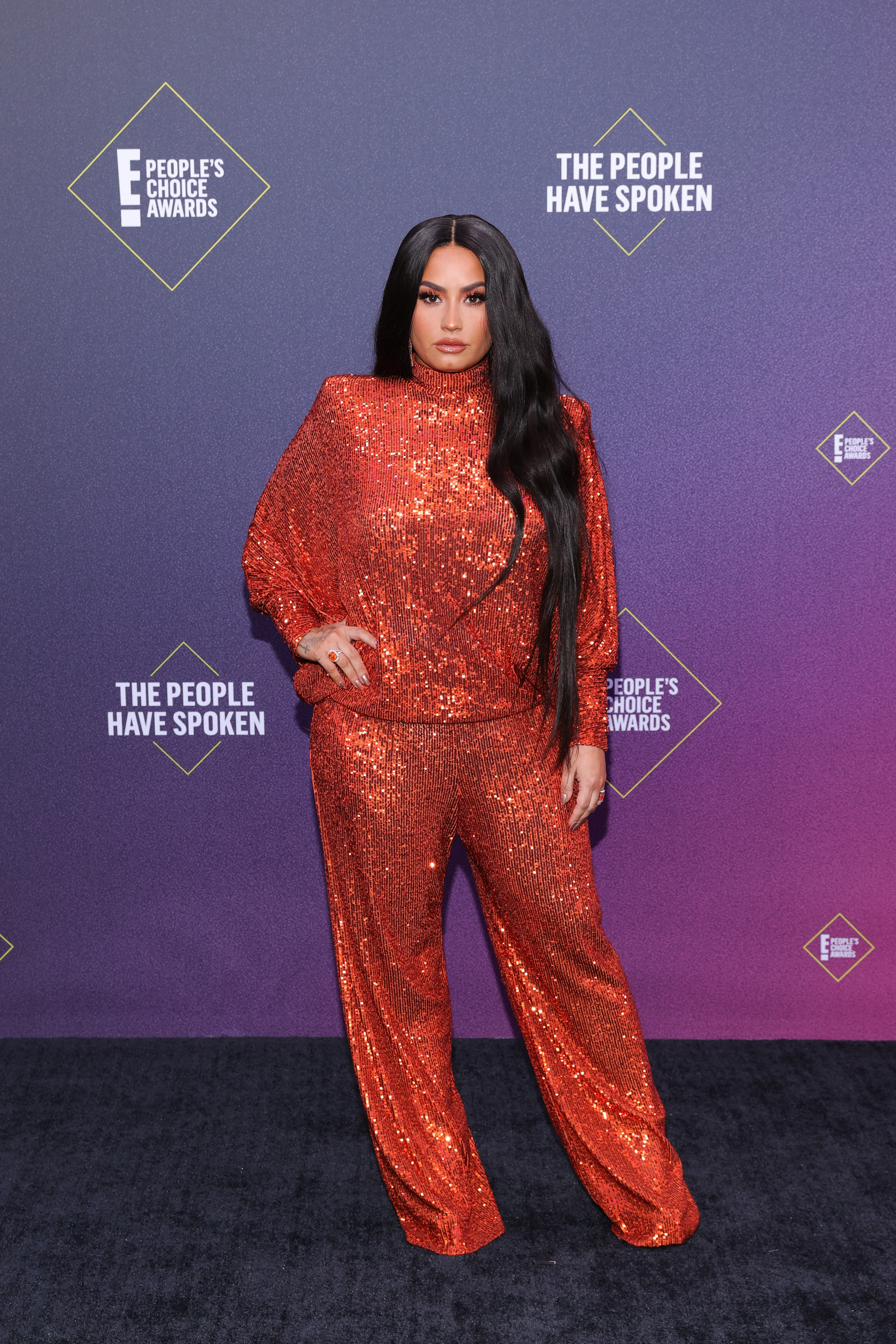 Demi Lovato poses in a shimmering red sequined ensemble at the E! People's Choice Awards in Santa Monica, California, exuding bold style on the event's purple carpet.