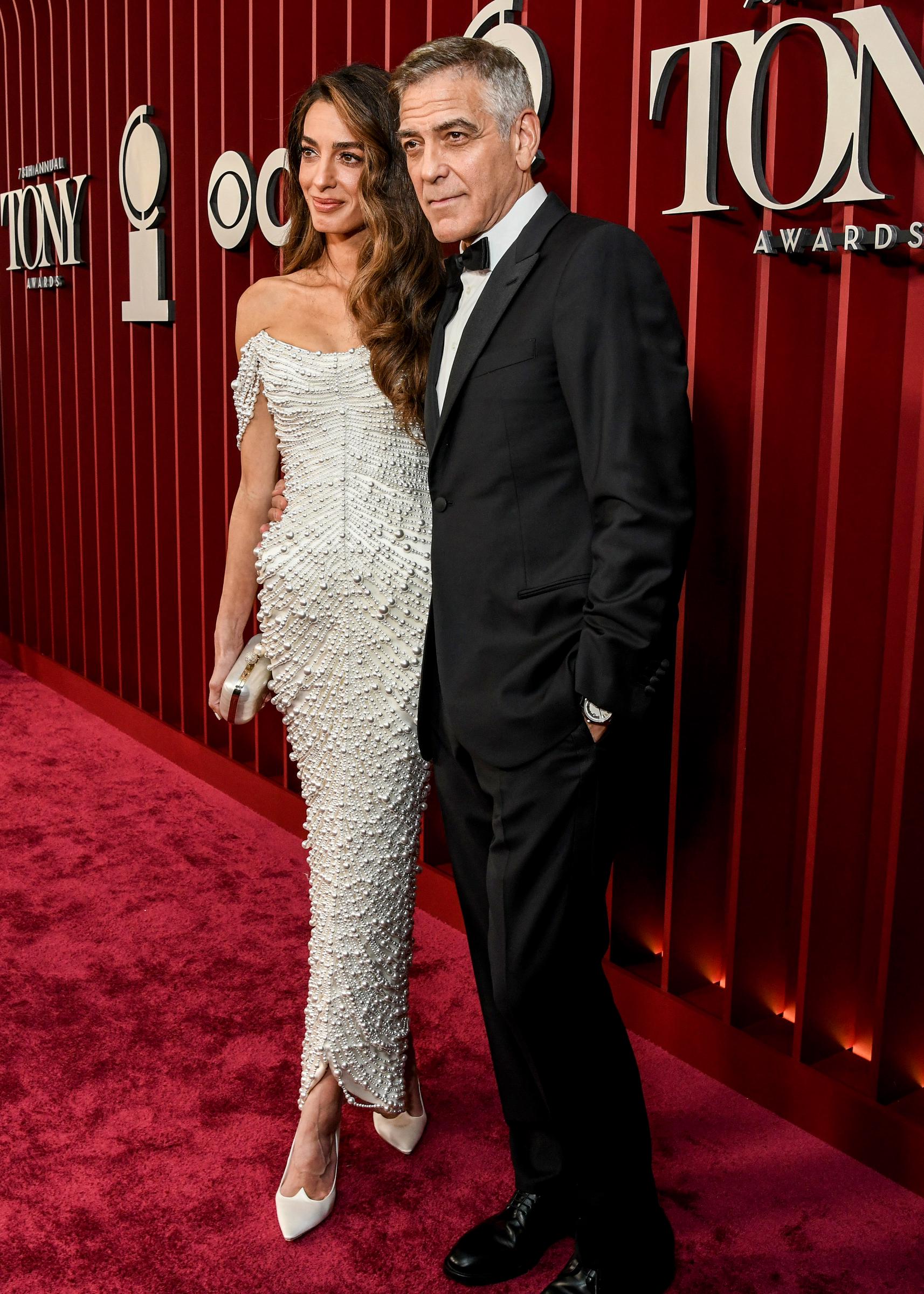 Amal Clooney stands with her feet crossed beside George Clooney at the Tony Awards. George appears relaxed as they pose together in front of the deep red backdrop.