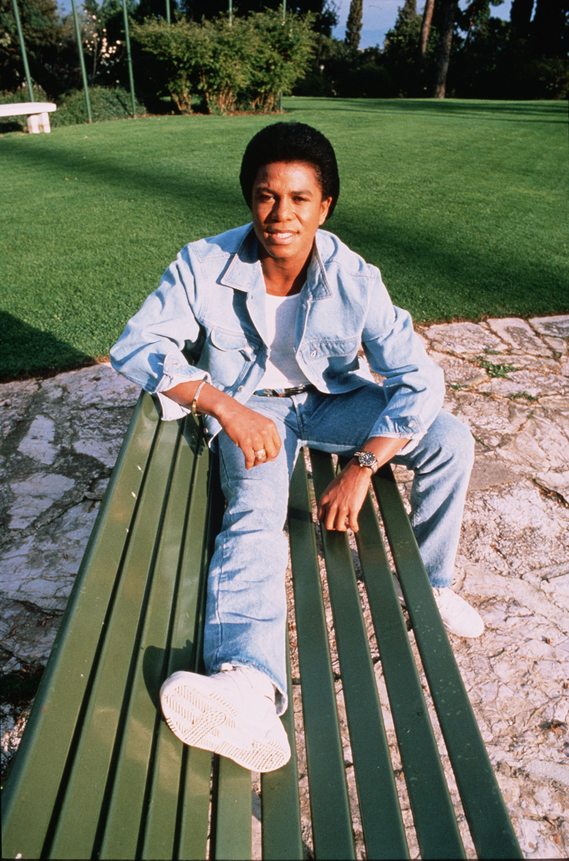 Seated casually on a green bench in Rome, Jermaine Jackson leans forward in a denim-on-denim look, blending effortless cool with a laid-back, off-duty vibe. The sunlit setting and relaxed posture add to the candid feel, offering a glimpse of the star away from the spotlight.