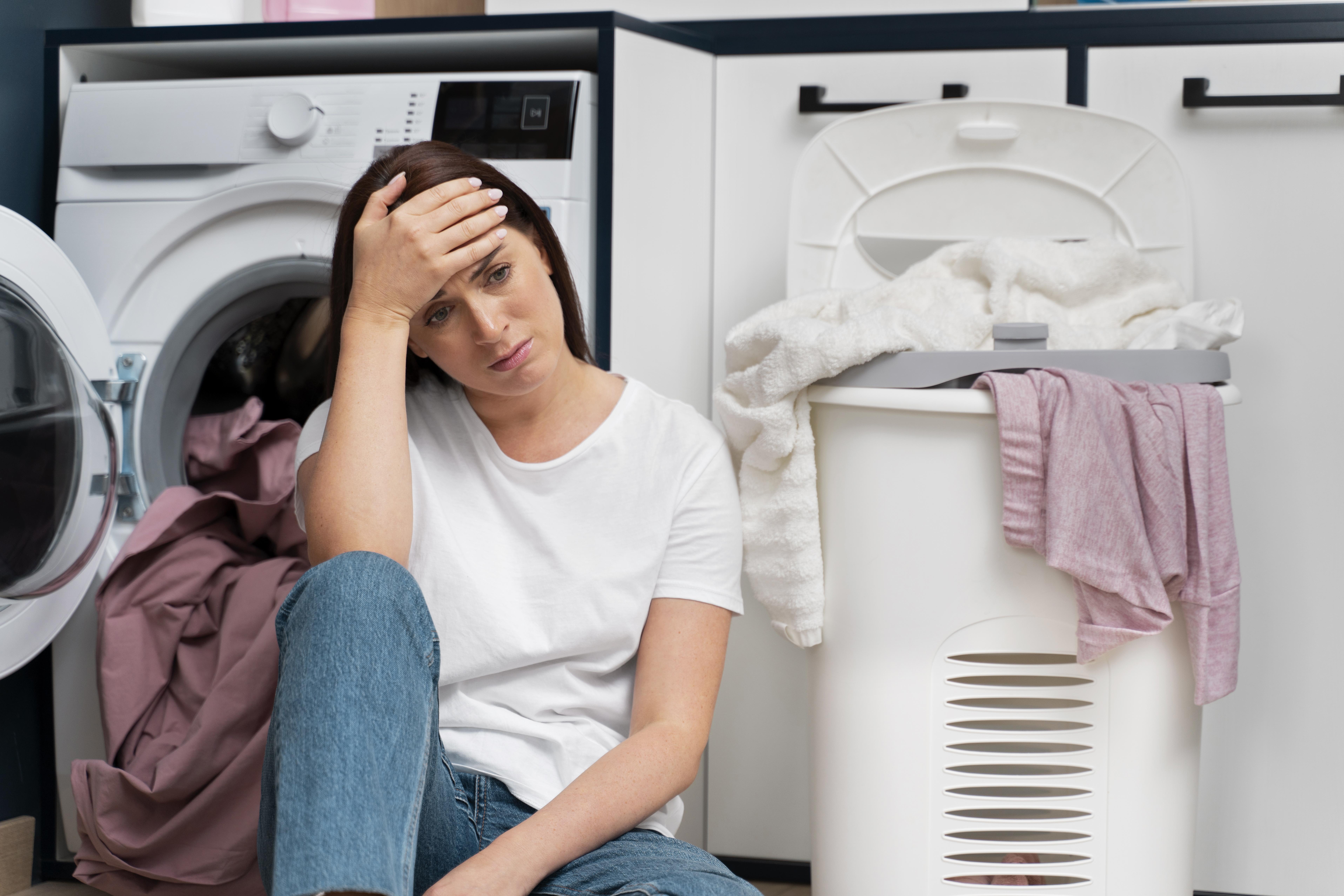 Woman looking upset as she's leans on a washing machine, surrounded by laundry | Source: Freepik