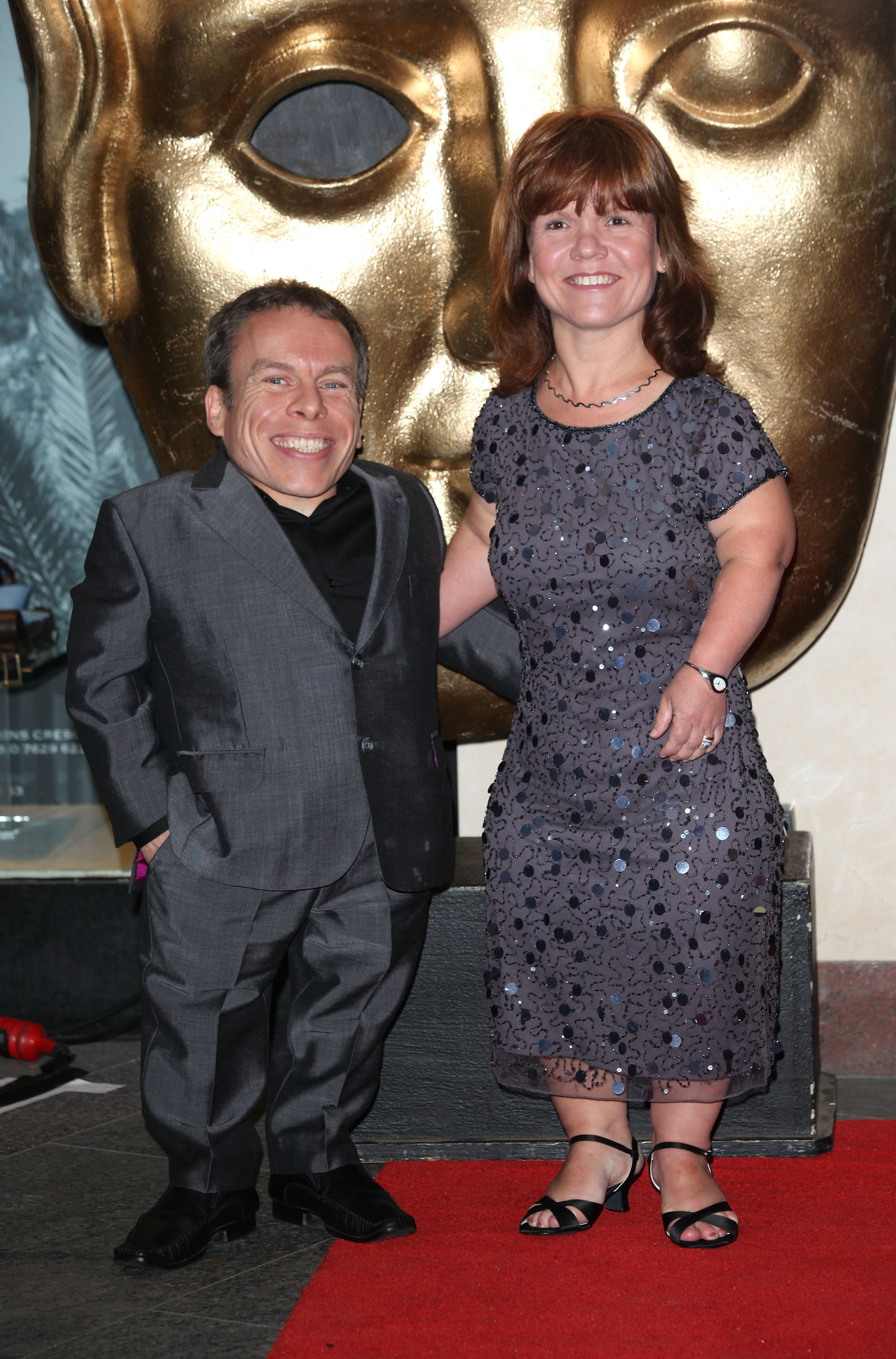 Warwick and Samantha Davis link arms on the red carpet at the British Academy Children's Awards in London, England, on 27 November 2011, both smiling broadly in front of the iconic golden BAFTA mask — a fitting backdrop for a couple who were no strangers to the industry's most prestigious stages. When Warwick received the BAFTA Fellowship years later, it was Samantha he chose to dedicate it to, describing it as "the best thing that's ever happened to me."