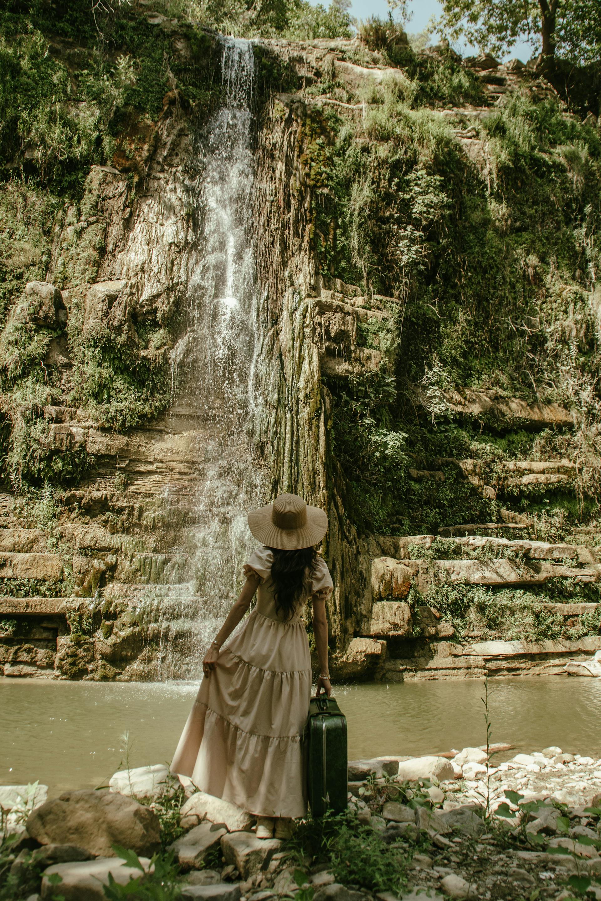 A woman in a beige dress and a straw hat looking at a waterfall | Source: Pexels