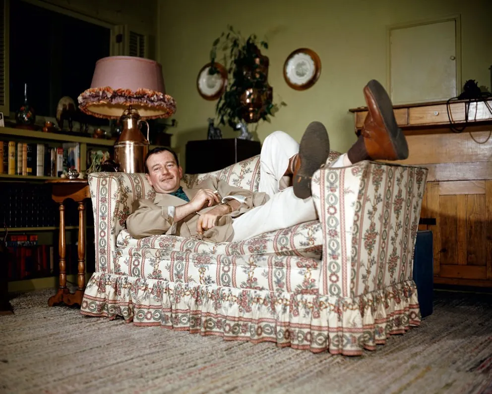 Portrait de l'acteur américain John Wayne (1907 - 1979), une cigarette à la main, alors qu'il se détend, les pieds sur l'accoudoir d'un canapé, dans les années 1950. | Source : Getty Images