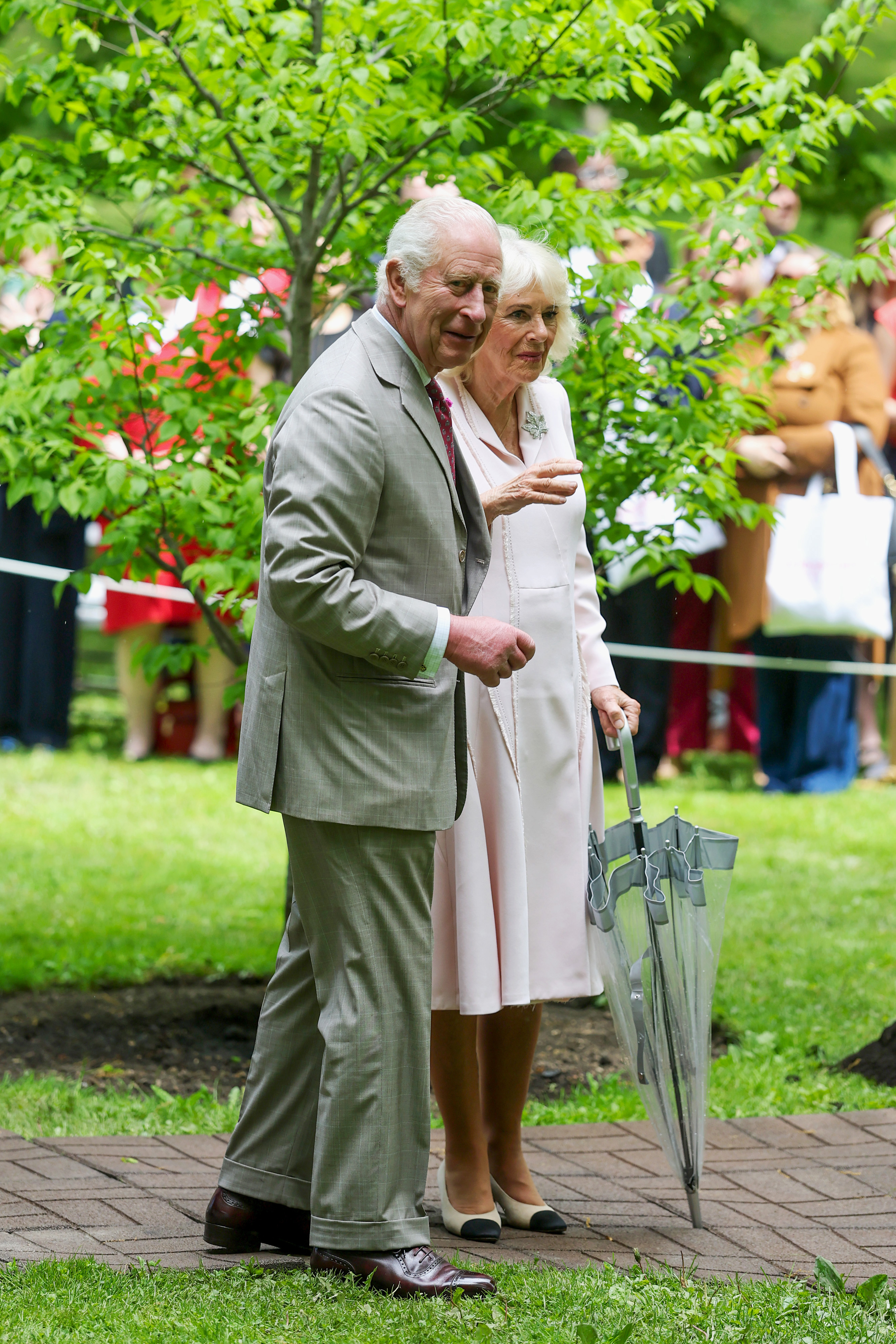 Camilla, with her closed umbrella in hand, gestures toward something in the distance as King Charles III, dressed in a grey suit for the occasion, looks on attentively.