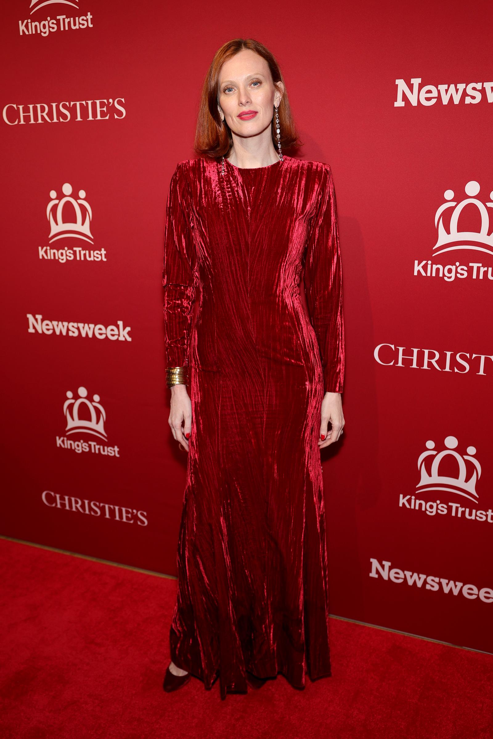 Karen Elson is head-to-toe in richly textured crushed crimson velvet, the full-length long-sleeved gown catching the light in shifting waves, set off by long dangling crystal earrings, a stacked gold cuff bracelet, red nails, and matching dark burgundy pointed-toe flats.