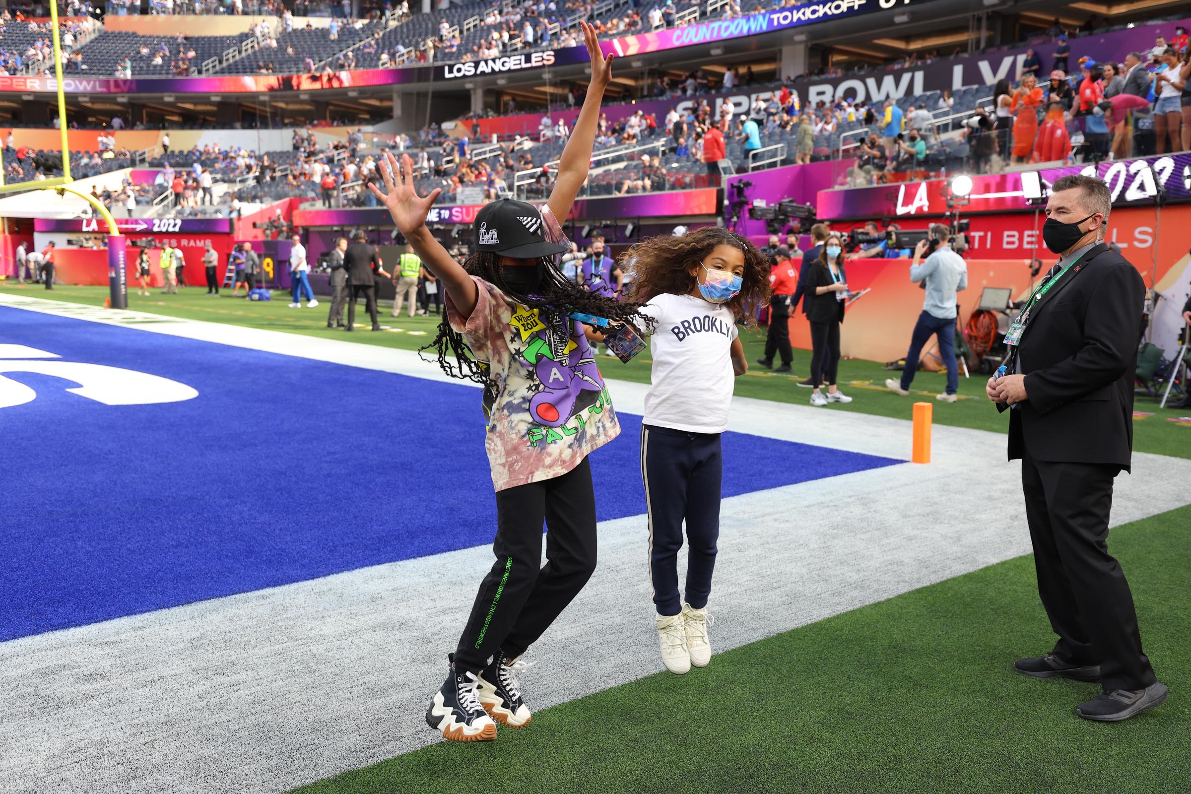 Blue Ivy jumps with her arms raised on the field before kickoff, her movement playful and energetic. The candid shot highlights her comfort and excitement in the Super Bowl setting.