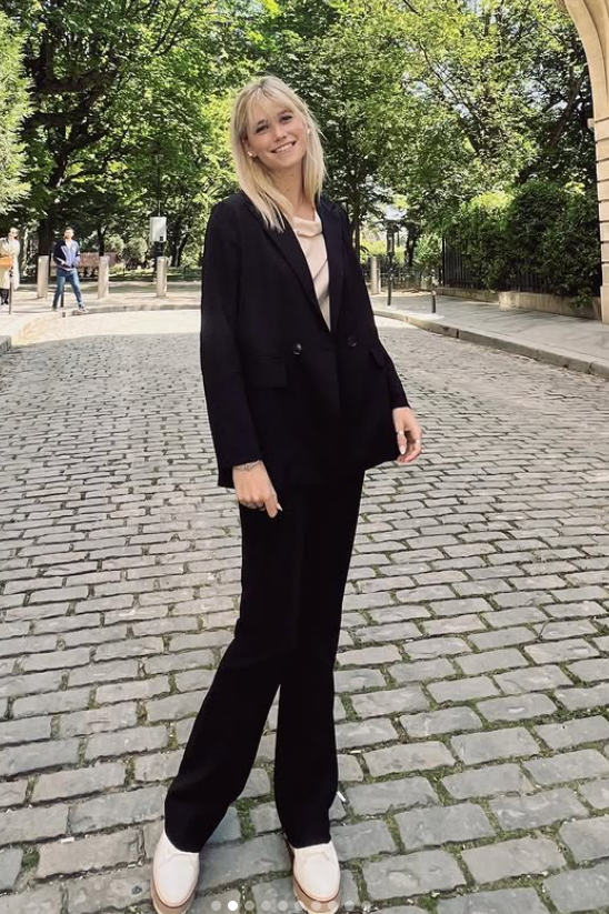 Effortlessly chic, Asleigh Scully smiles on a cobblestone street, her relaxed black suit and soft posture capturing a blend of elegance and easy confidence in a leafy city setting. | Source: Instagram/ascullyphoto