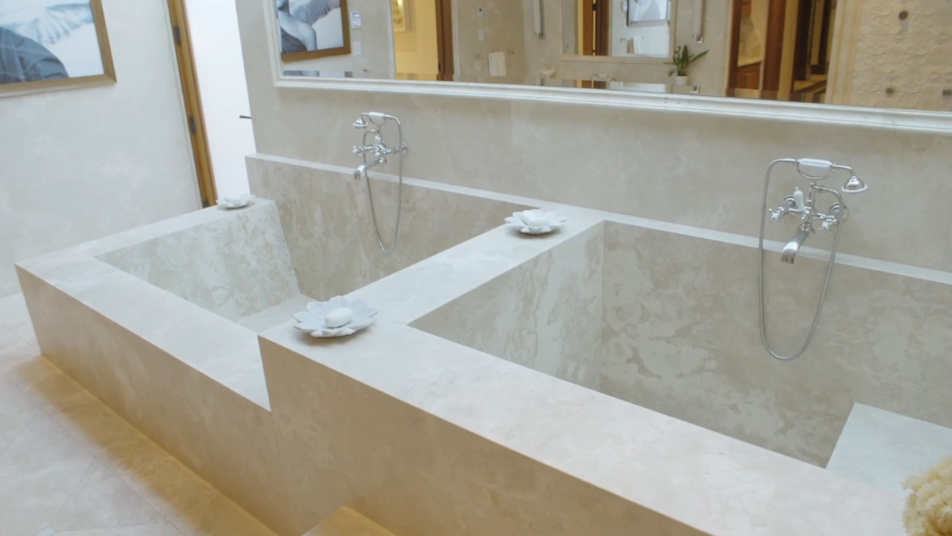 Twin custom-built stone soaking tubs finished in smooth beige travertine sit side-by-side in this luxury spa bathroom. Polished chrome telephone-style faucets and flower-shaped soap dishes complement the minimalist, resort-inspired bath sanctuary. | Source: YouTube/Architectural Digest