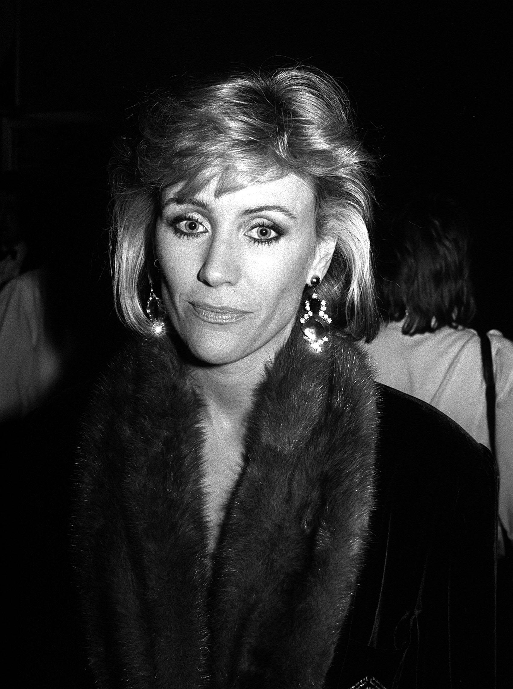 Angie Best is pictured at an evening event, wearing statement earrings and a dark outfit with a fur-trimmed collar, her softly styled blonde hair and direct gaze capturing the polished glamour of the era.