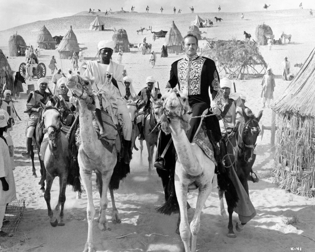 Charlton Heston and Johnny Sekka riding a camel with a group of men behind him riding horseback in a scene from the film 'Khartoum', 1966. | Photo: Getty Images