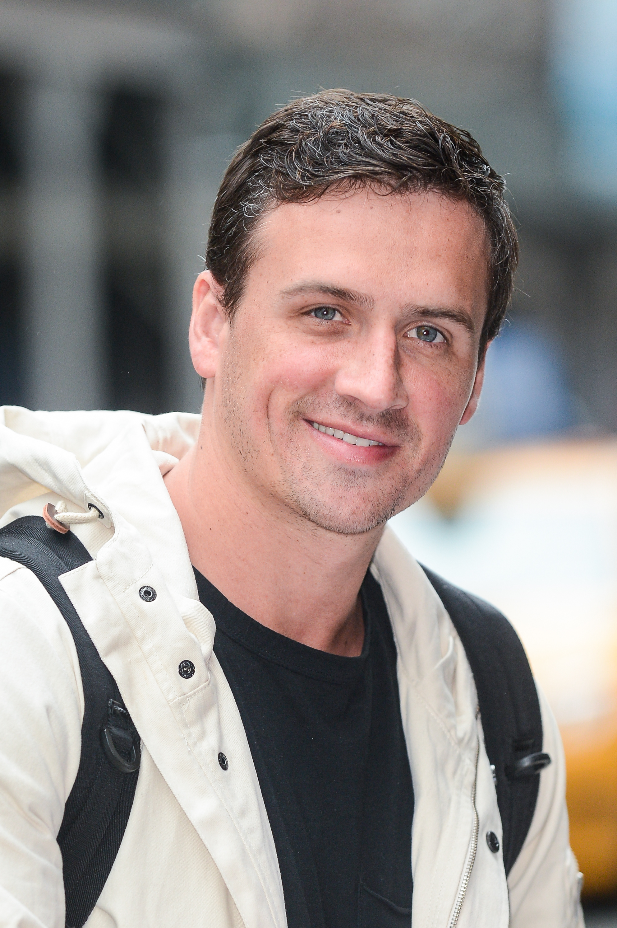 Ryan Lochte smiles while leaving his Tribeca hotel in New York City, wearing a light-colored jacket layered over a black shirt. The Olympic swimmer carries a backpack over both shoulders as he walks through the busy street entrance.