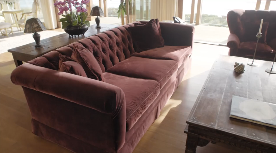 A plush tufted velvet sofa in a rich plum tone anchors a cozy sitting area within the primary suite, illuminated by soft natural sunlight. An antique-style wooden coffee table and a console table decorated with vibrant potted orchids sit nearby, enhancing the space's elegant character. | Source: YouTube/Architectural Digest