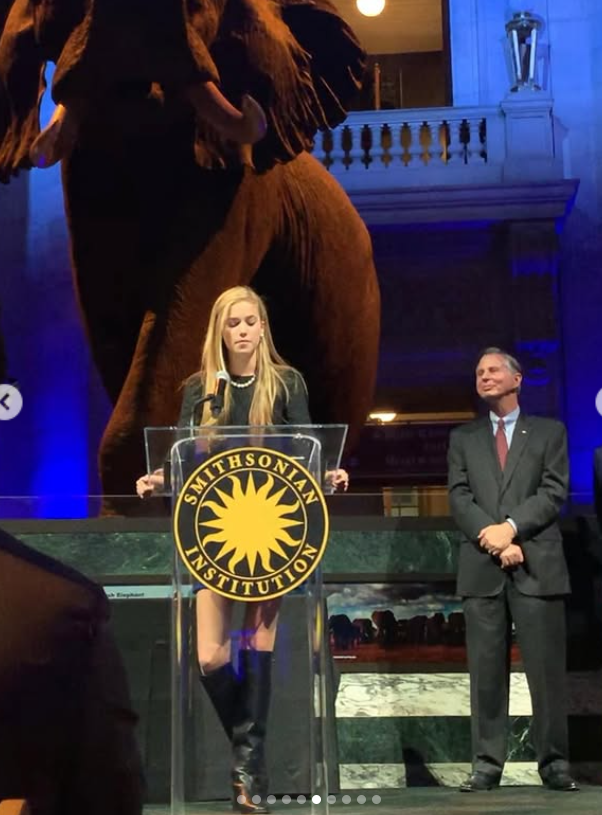 Standing at a podium, Asleigh Scully delivers remarks beneath towering museum displays, the setting adding weight to her composed presence. | Source: Instagram/ascullyphoto