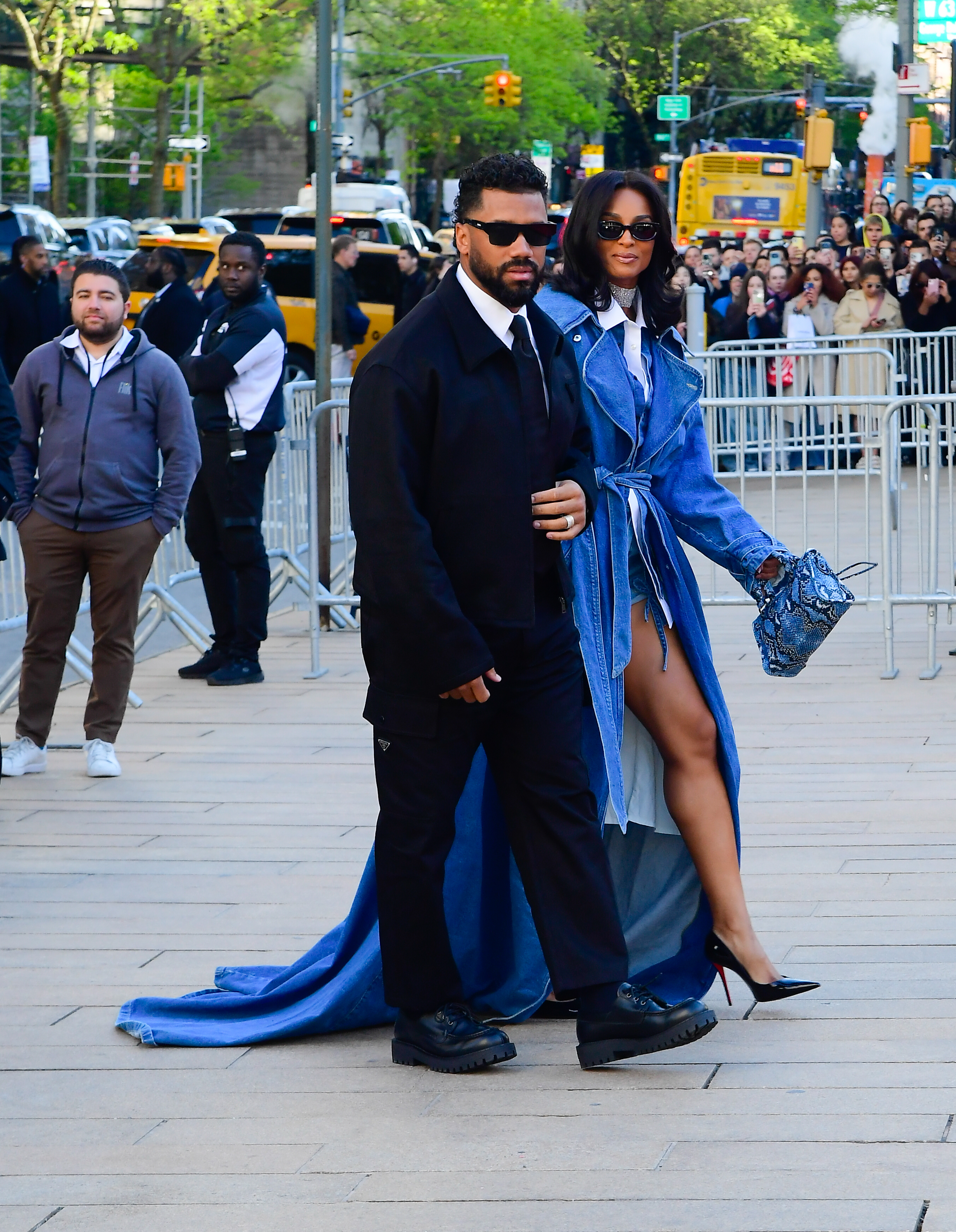 Russell Wilson and Ciara walk past a crowd of onlookers, maintaining a cool, composed presence amid the busy scene. Both wear dark sunglasses, adding a touch of mystery as her long coat flows behind her.