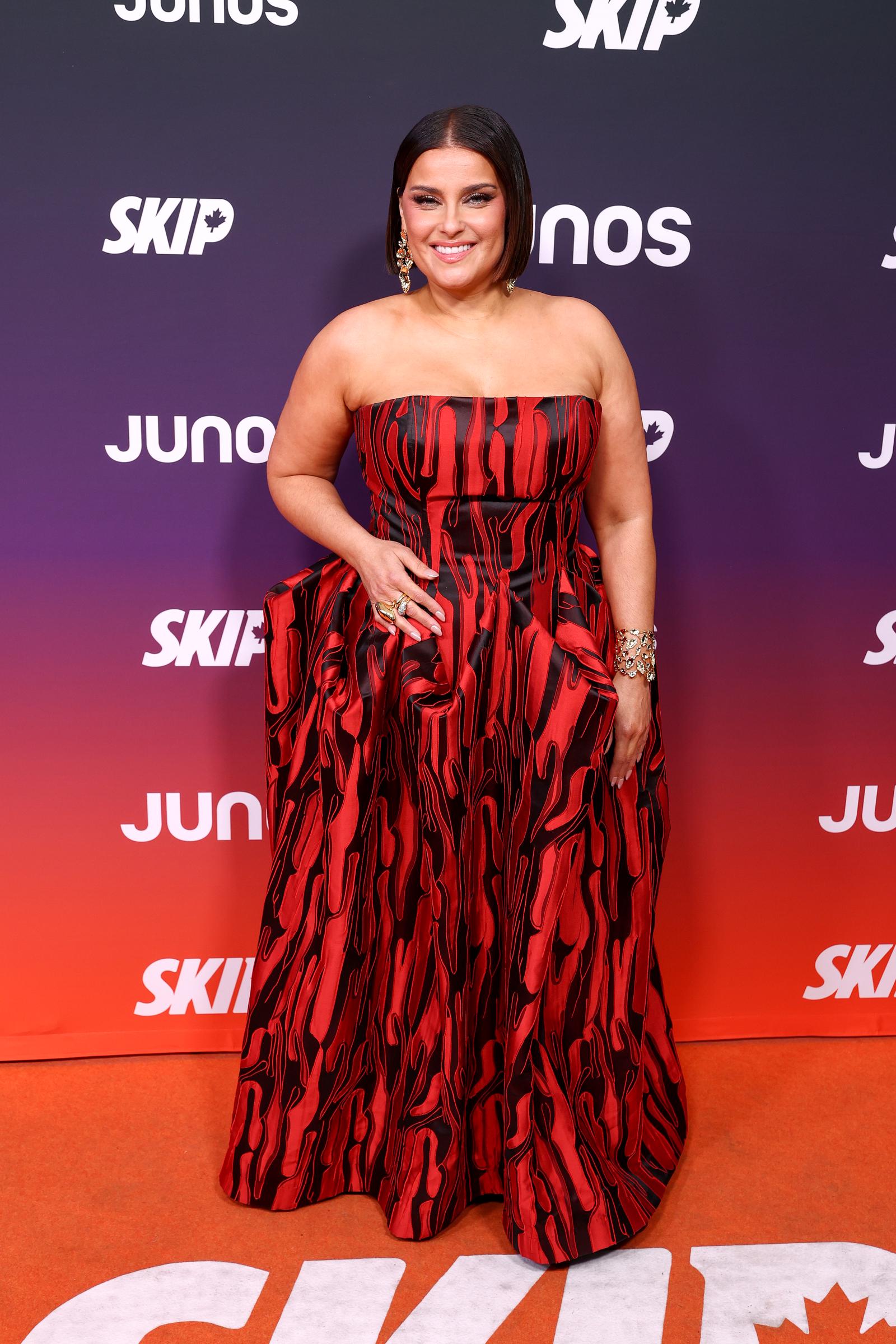 Nelly Furtado shines at the JUNO Awards in Hamilton, Ontario, wearing a striking strapless red-and-black gown with a bold pattern, posing confidently on the red carpet with elegant jewelry and a radiant smile.