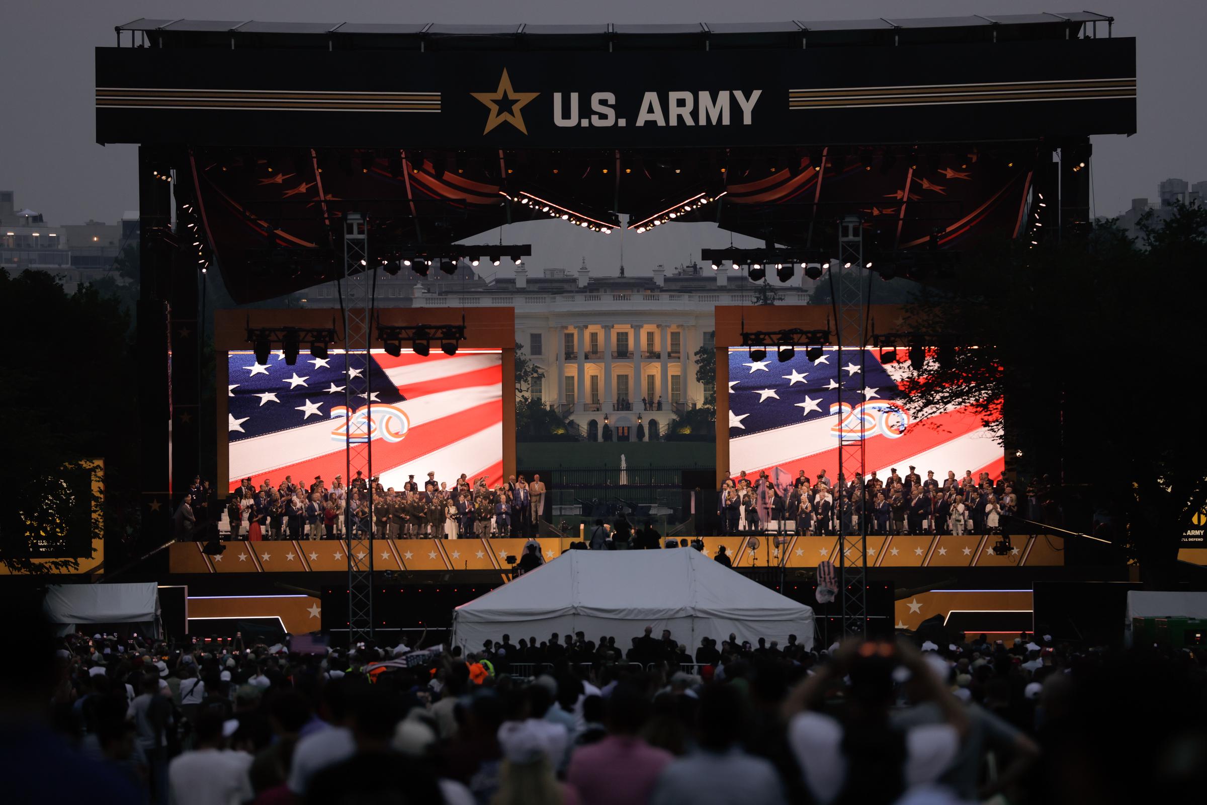As the sun sets, a large crowd watches the US Army’s 250th anniversary ceremony unfold. The stage is lit with giant American flags and a view of the White House in the distance, emphasizing the event’s national scale and ceremonial pride.