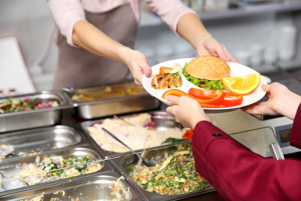 Mujer agradable dando almuerzo a una colegiala en la cafetería escolar. | Fuente: Shutterstock. Mujer agradable dando almuerzo a una colegiala en la cafetería escolar. | Fuente: Shutterstock.