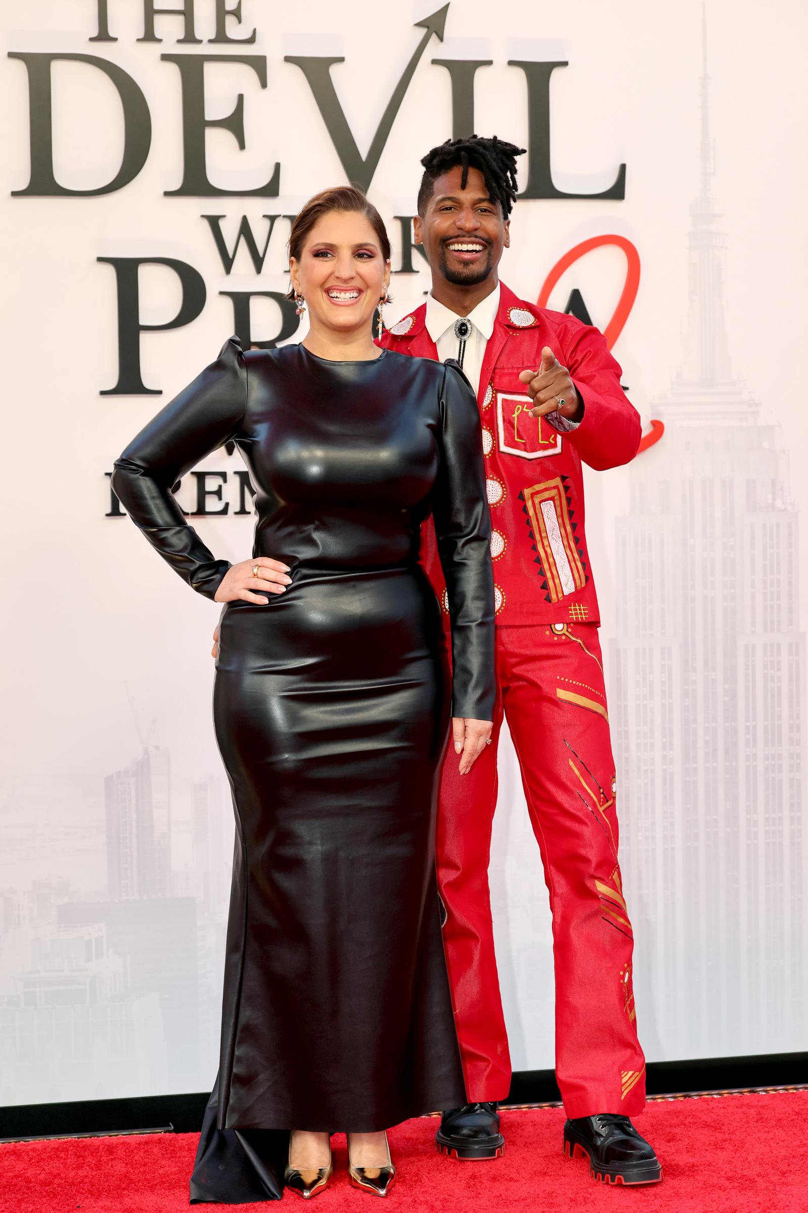 Suleika Jaouad wears a long-sleeved black leather column gown with gold pointed-toe heels and chandelier earrings, while her husband Jon Batiste steals the scene in a head-to-toe red embroidered suit featuring geometric and circular motifs, a white dress shirt, black bolo tie, and chunky black platform shoes.