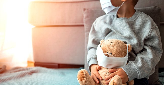 A young child wearing a mask holding a teddy bear. | Photo: Shutterstock