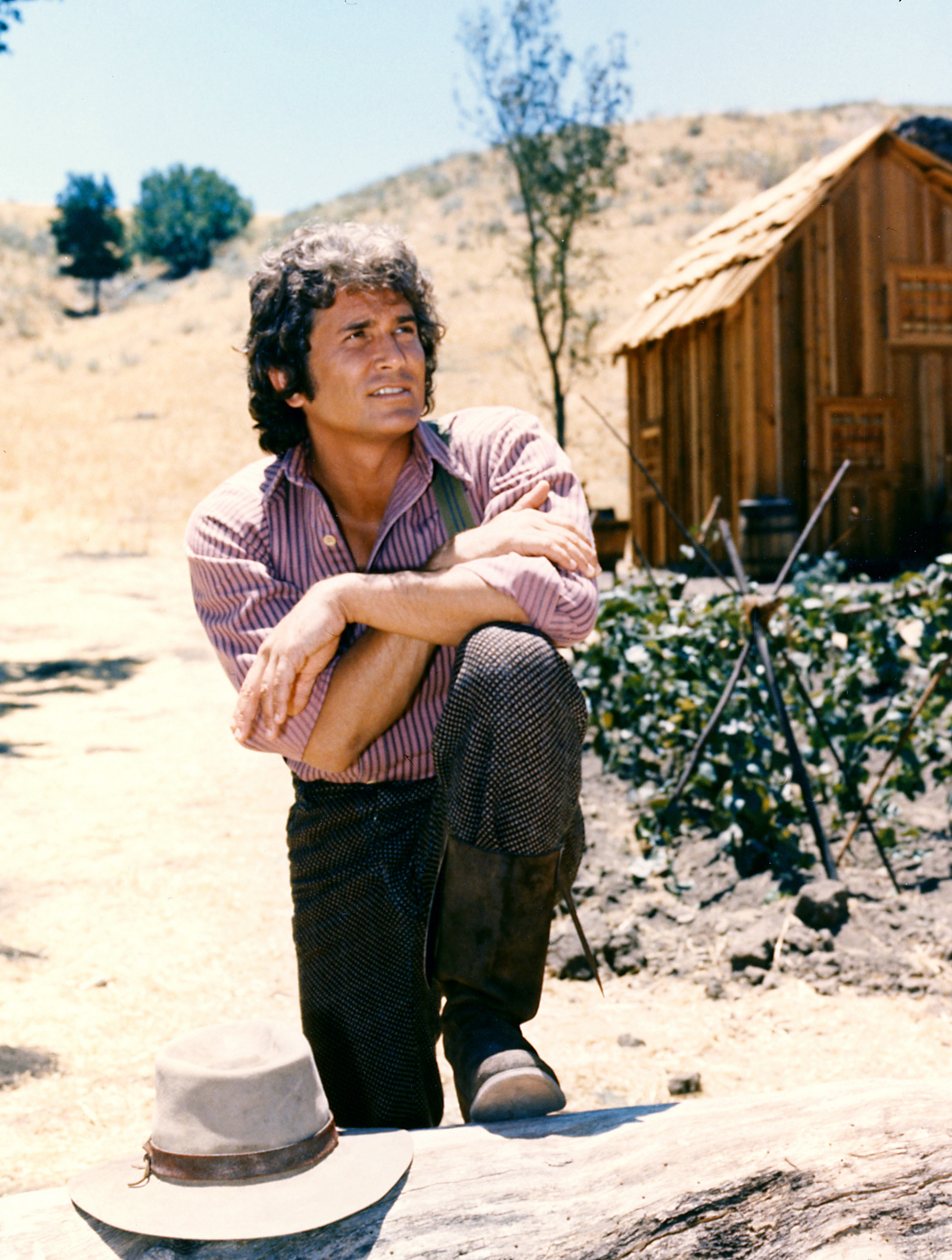 Michael Landon poses with his arms crossed and resting on his knee with a log cabin in the background in a portrait for the US television series, 'Little House on the Prairie,' circa 1974. | Source: Getty Images