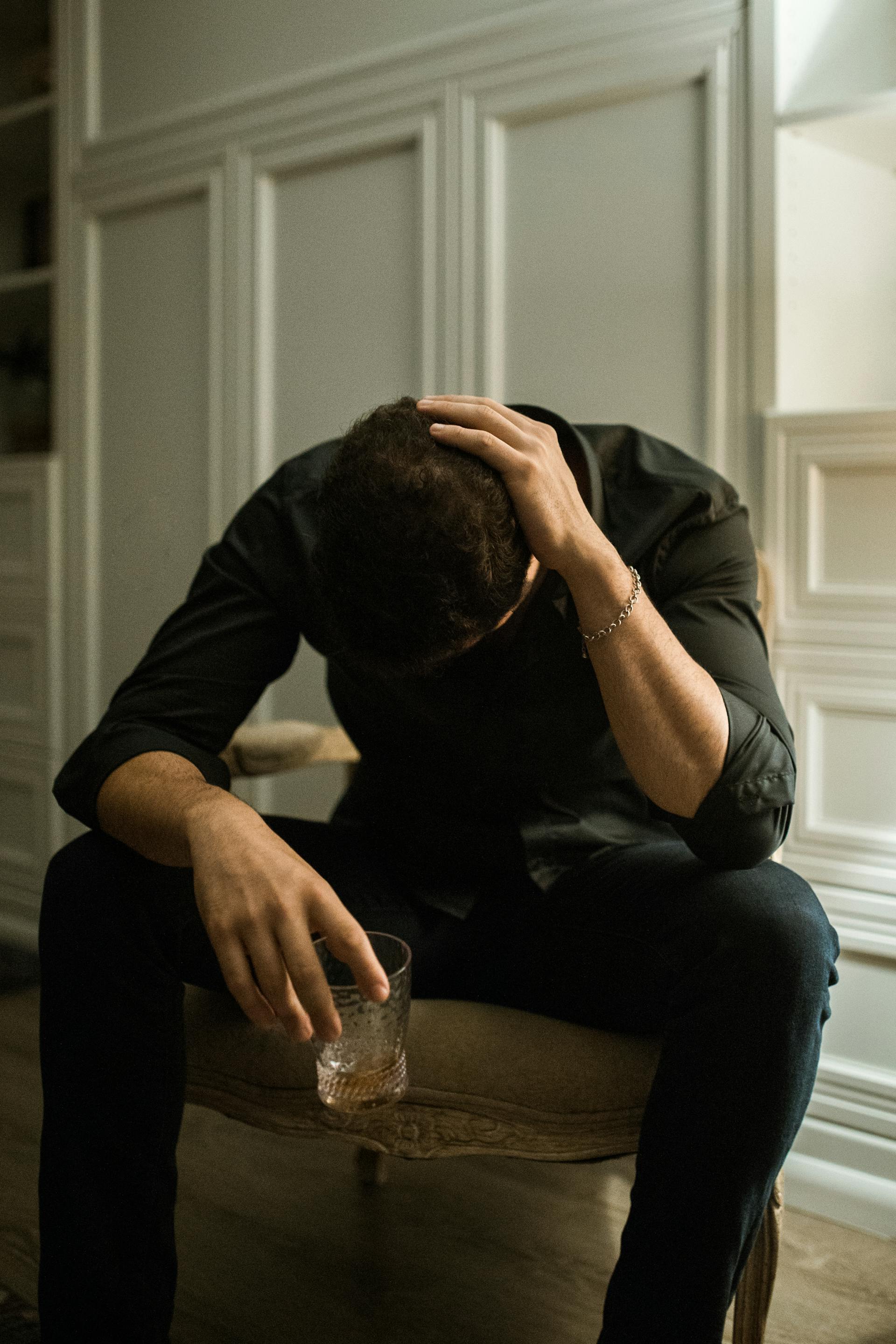 A depressed man sitting on a chair with his head bowed while holding a glass of drink | Source: Pexels