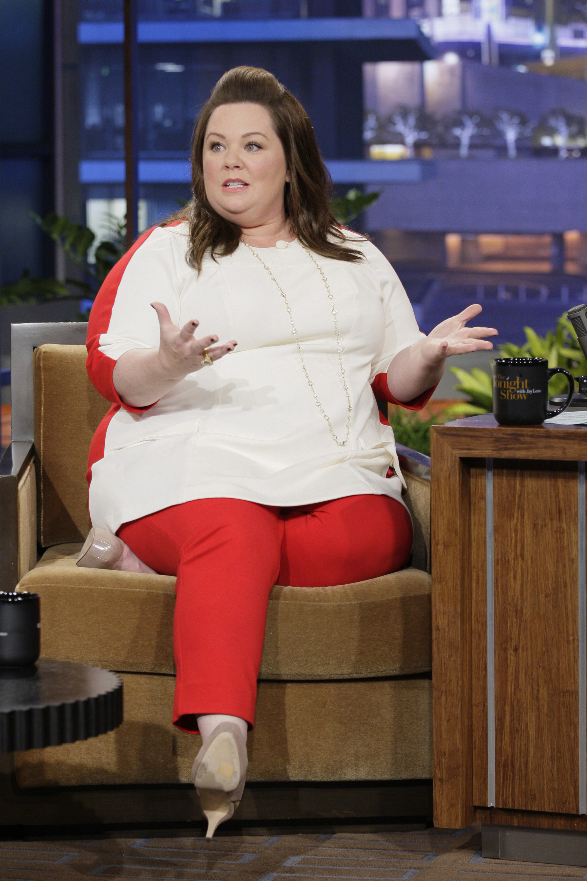 Melissa McCarthy sits animatedly in the interview chair, dressed in a crisp white top and bold red pants that energize the scene. With expressive gestures and a lively presence, she brings humor and warmth to the late-night set.