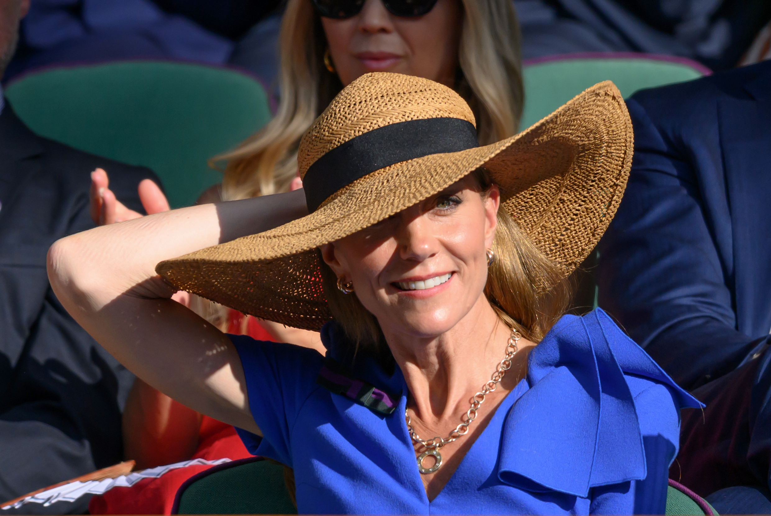 Princess Catherine smiles warmly as she lifts a hand to adjust her hat during the match. Sunlight filters through the brim, catching the angles of her face.