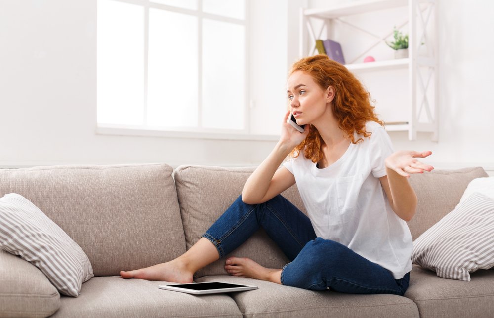 A young agitated redhead woman sitting on a couch while talking on the phone | Photo: Shutterstock/Prostock-studio