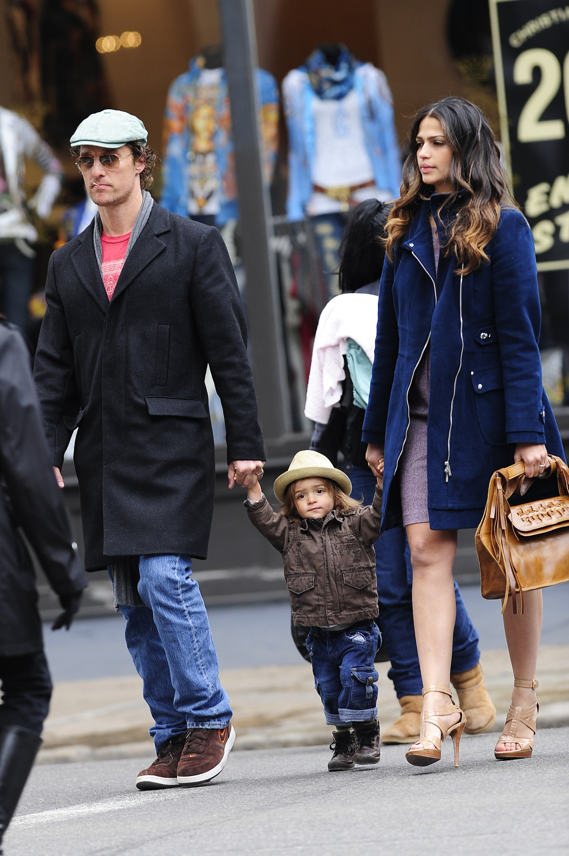 Levi McConaughey walks between Matthew McConaughey and Camila Alves, holding both their hands. He wears a hat, jacket, and pants while stepping forward. Matthew McConaughey and Camila Alves walk on either side in coats and sunglasses.