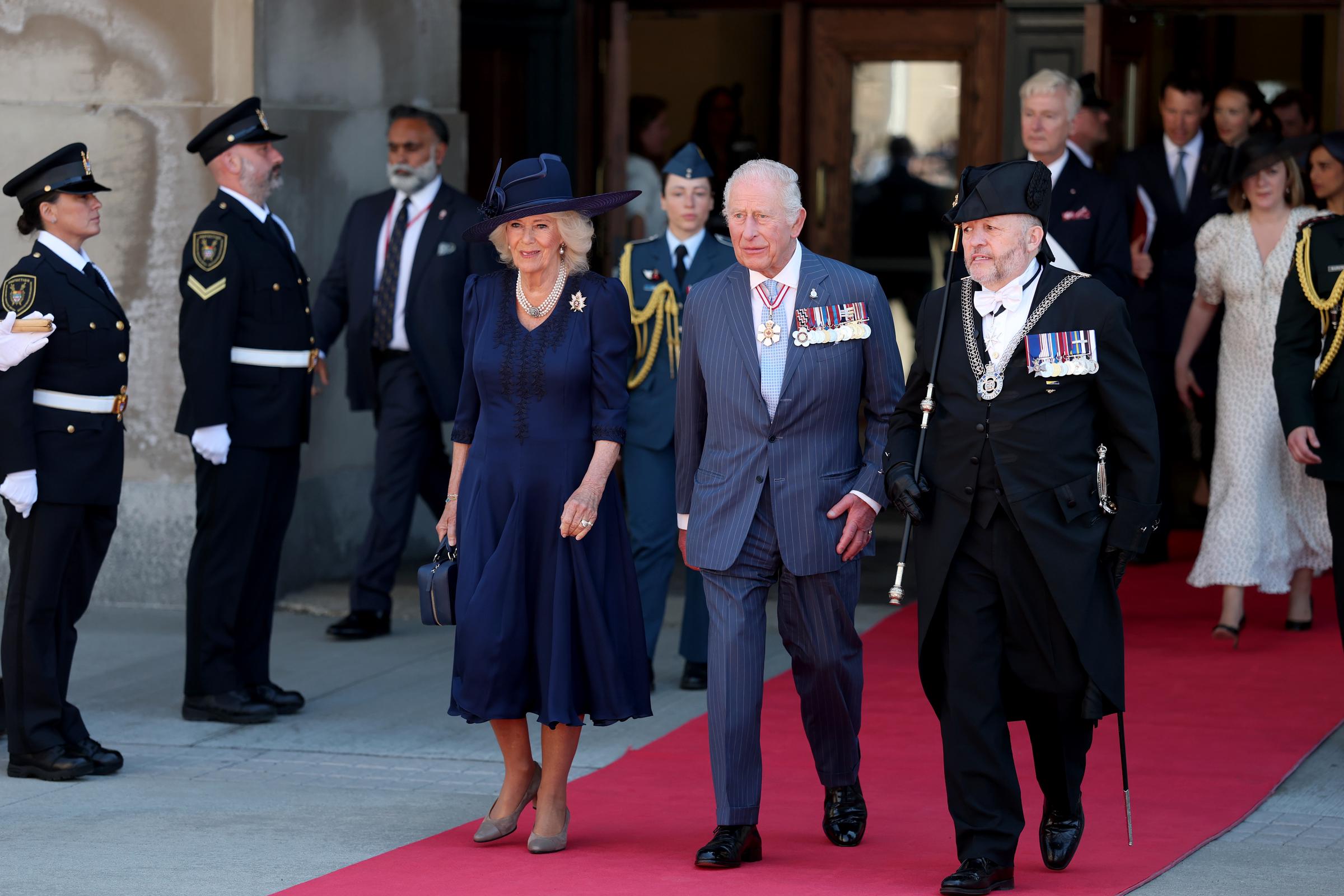 King Charles III and Queen Camilla leave the Senate of Canada Building, with Camilla smiling warmly and Charles engaged in conversation with an official. The visit marks their first to Canada since 2022.