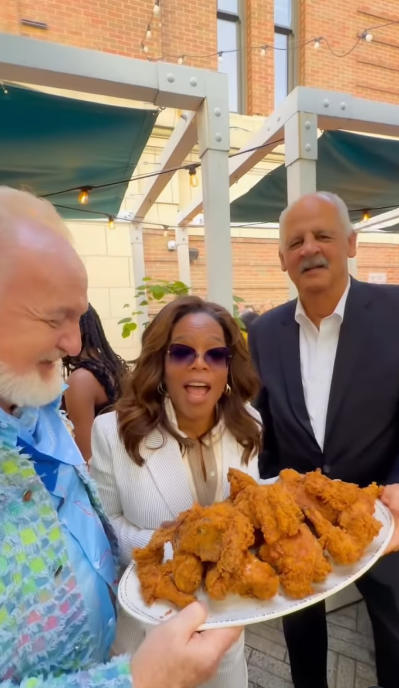 Oprah Winfrey appears animated beside Stedman Graham, smiling with her mouth open as they gather around a platter of fried chicken. Graham stands close by with a calmer, more reserved expression. | Source: Instagram/oprah