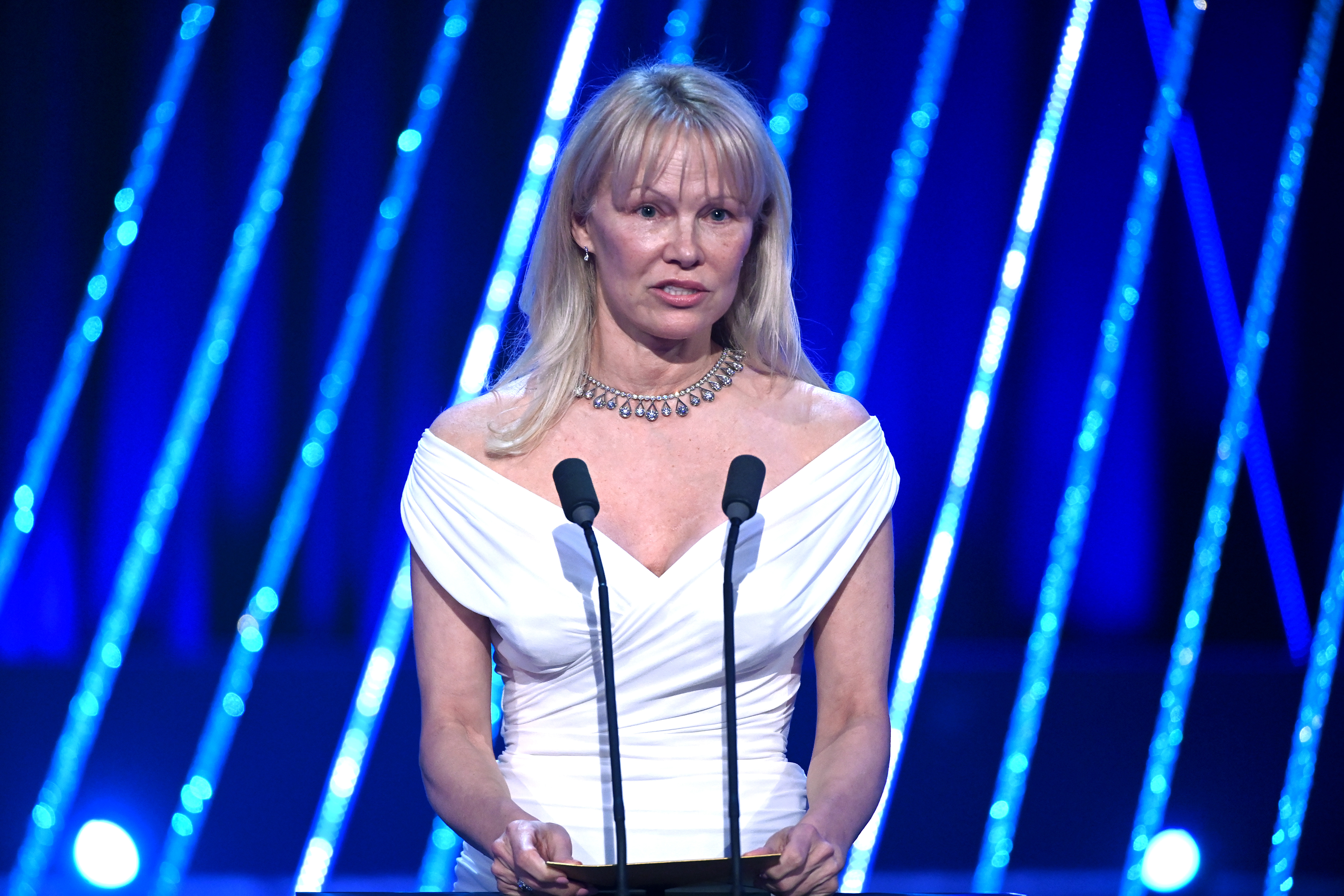 In a medium shot from the EE BAFTA Film Awards, Pamela Anderson presents the Leading Actor Award in a crisp white dress that radiates elegance. Her front bangs frame her face softly, complemented by minimal makeup for a natural glow. A delicate necklace and matching earrings add a touch of sophistication to her poised stage presence.