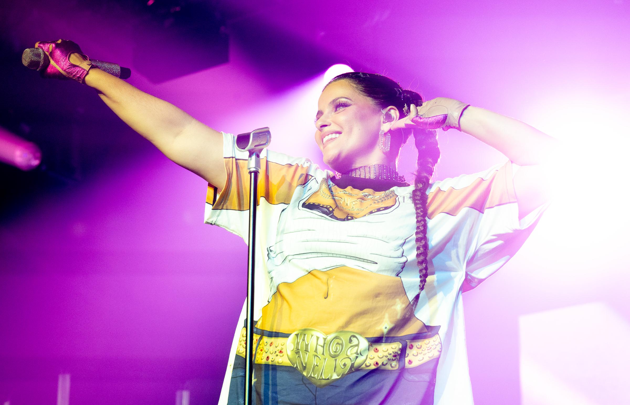 Furtado performs in an oversized cartoon-printed T-shirt featuring a crop top and denim mini skirt design, with a “Whoa Nelly” belt graphic. She wears bright fingerless gloves and braided hair, standing in pink stage lighting with a wide smile.