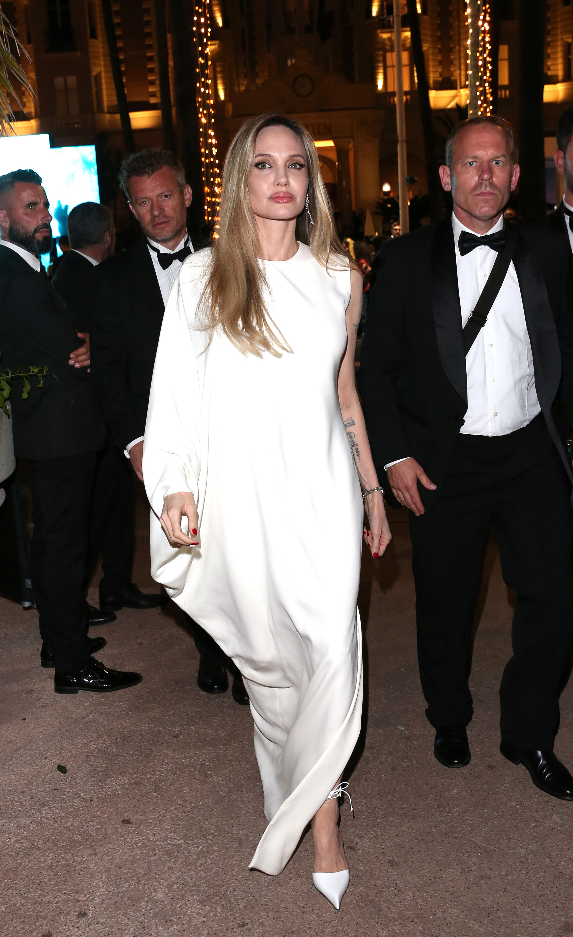 Angelina Jolie is seen arriving at the Chopard Trophy photocall in Cannes, France, flanked by security while wearing a flowing white gown and pointed heels.