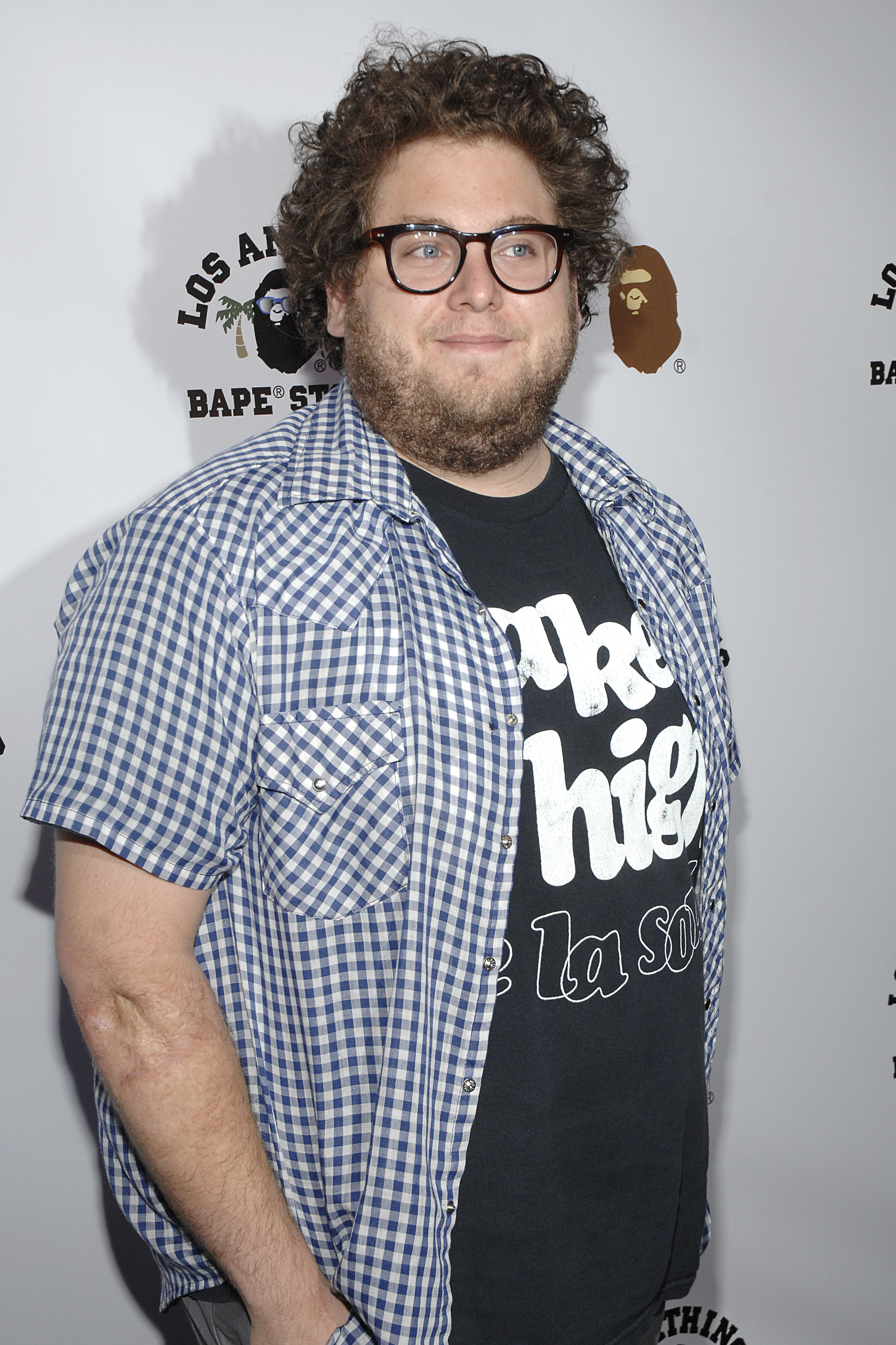 Jonah Hill attends a Los Angeles event in a blue-and-white checkered short-sleeve shirt layered over a graphic tee. With tousled curls, thick-rimmed glasses, and a full beard, the actor poses casually on the red carpet.