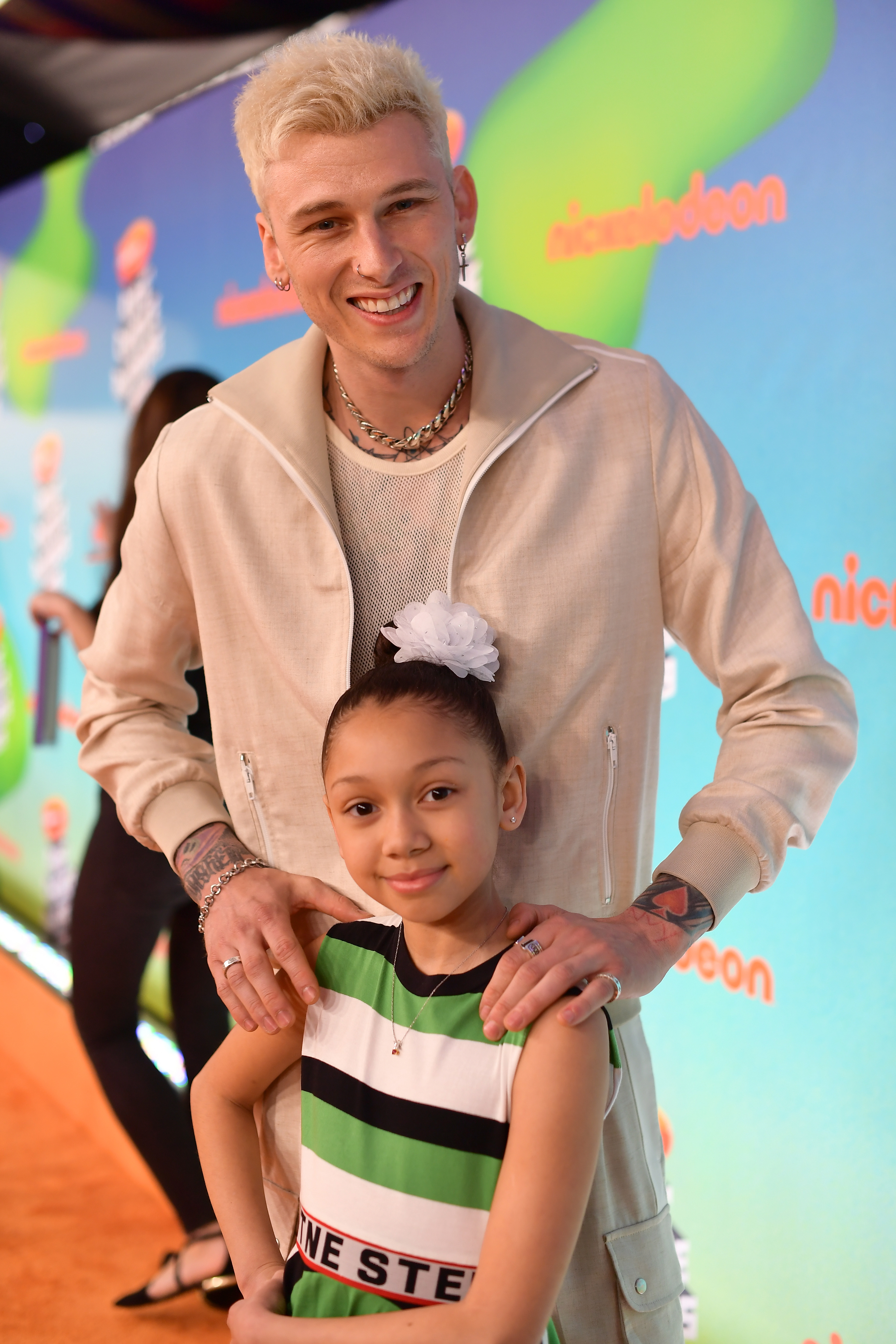 Machine Gun Kelly stands behind Casie Baker with his hands gently on her shoulders at the 2019 Kids' Choice Awards. Casie poses with confidence in a striped dress and white bow, smiling sweetly.