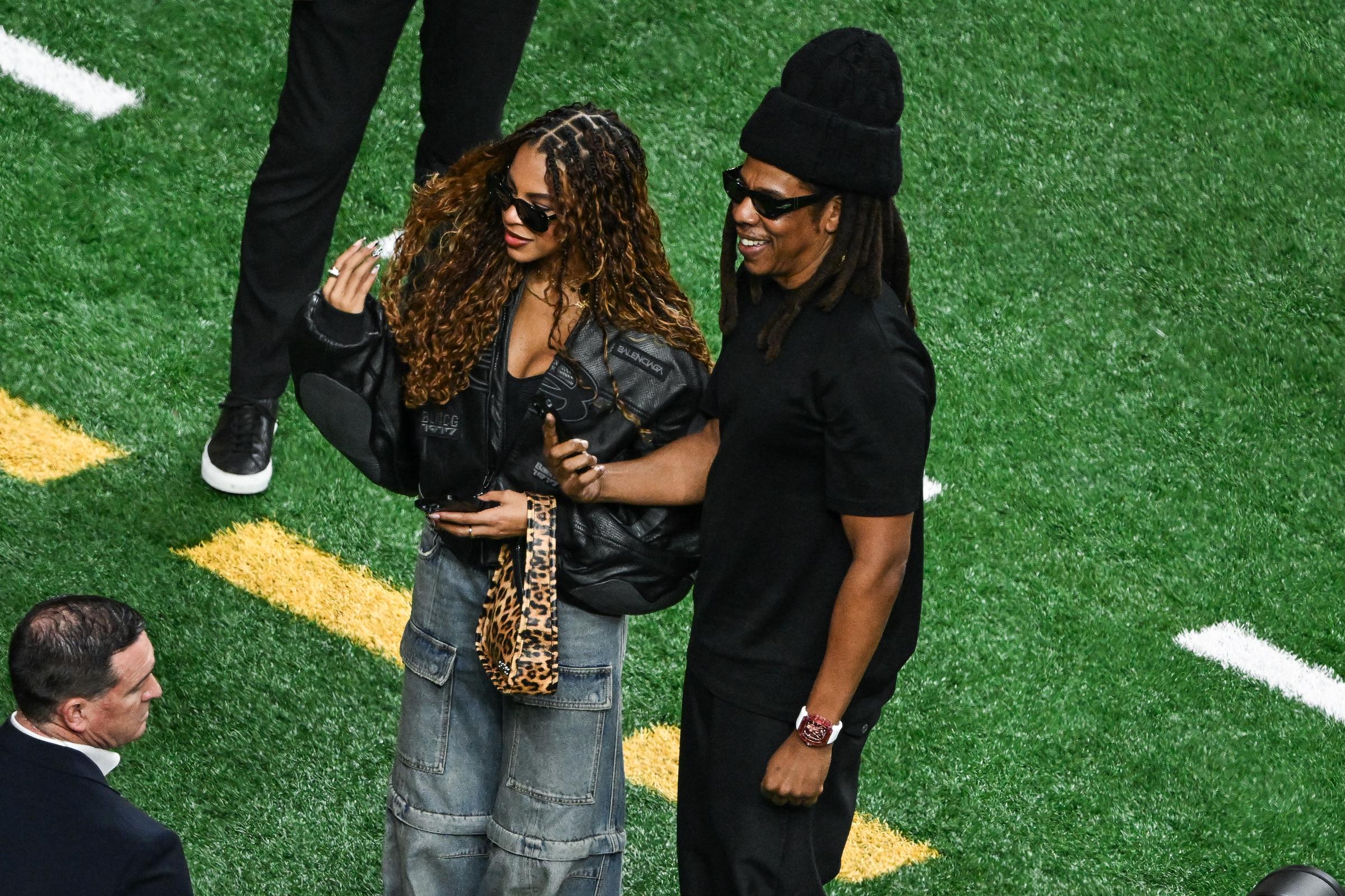 Blue Ivy stands close to Jay-Z, lifting her hand mid-gesture as he smiles beside her. Her loose curls and layered outfit contrast with his all-black look in the candid, side-by-side moment.