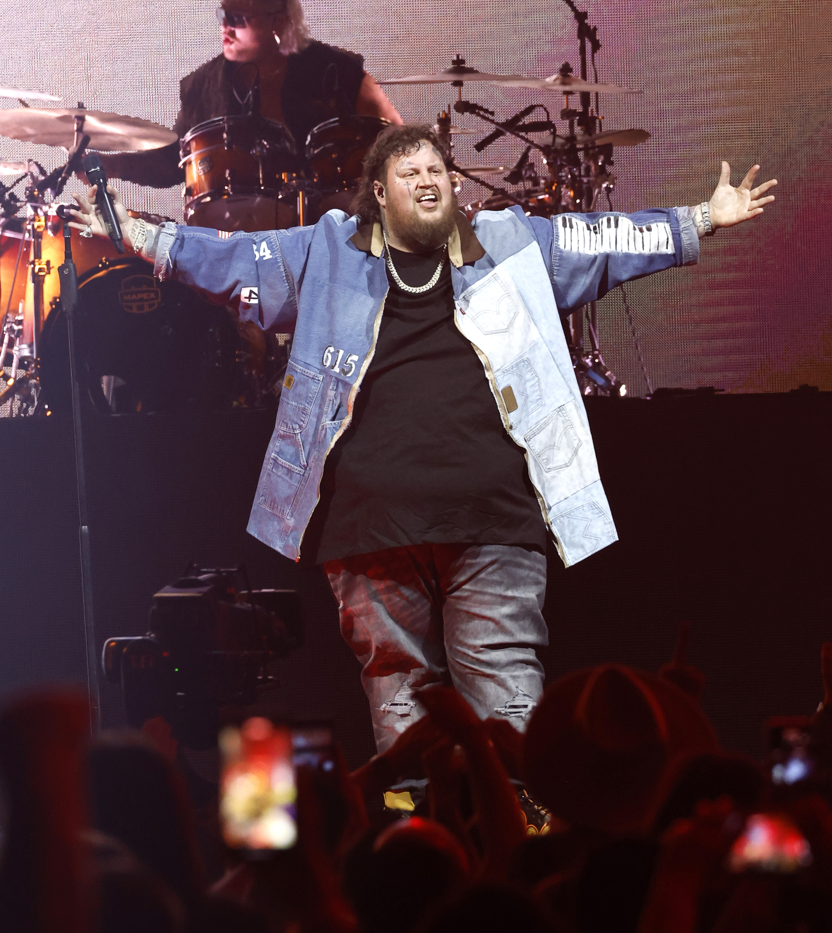 Jason "Jelly Roll" DeFord stands at center stage with arms wide open, exuding confidence and charisma as he engages with the crowd. He wears a patchwork denim jacket over a black shirt and distressed jeans, accented with a thick silver chain. His expression radiates energy and triumph, capturing the electric atmosphere of his live performance.