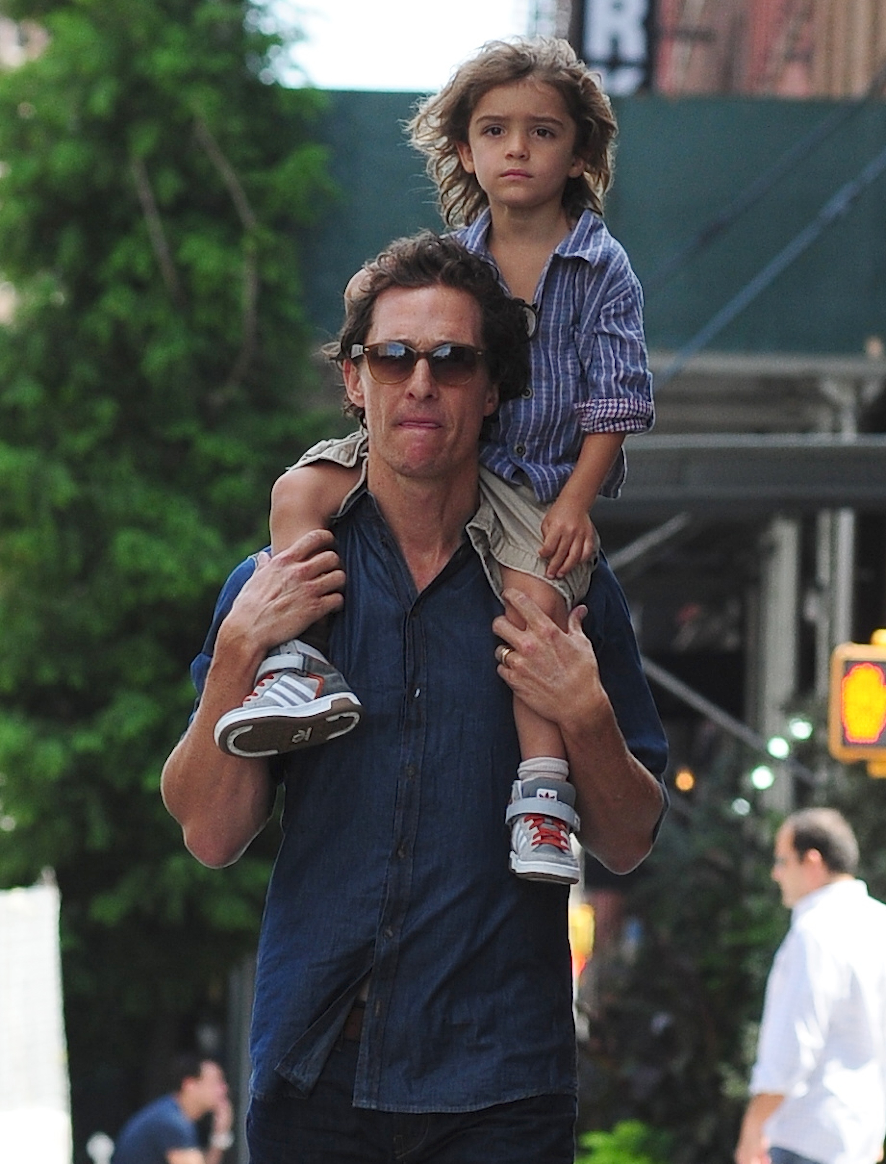 Levi Alves McConaughey sits on Matthew McConaughey’s shoulders. He looks ahead while holding steady as they walk. Matthew McConaughey supports his legs and moves along the street.