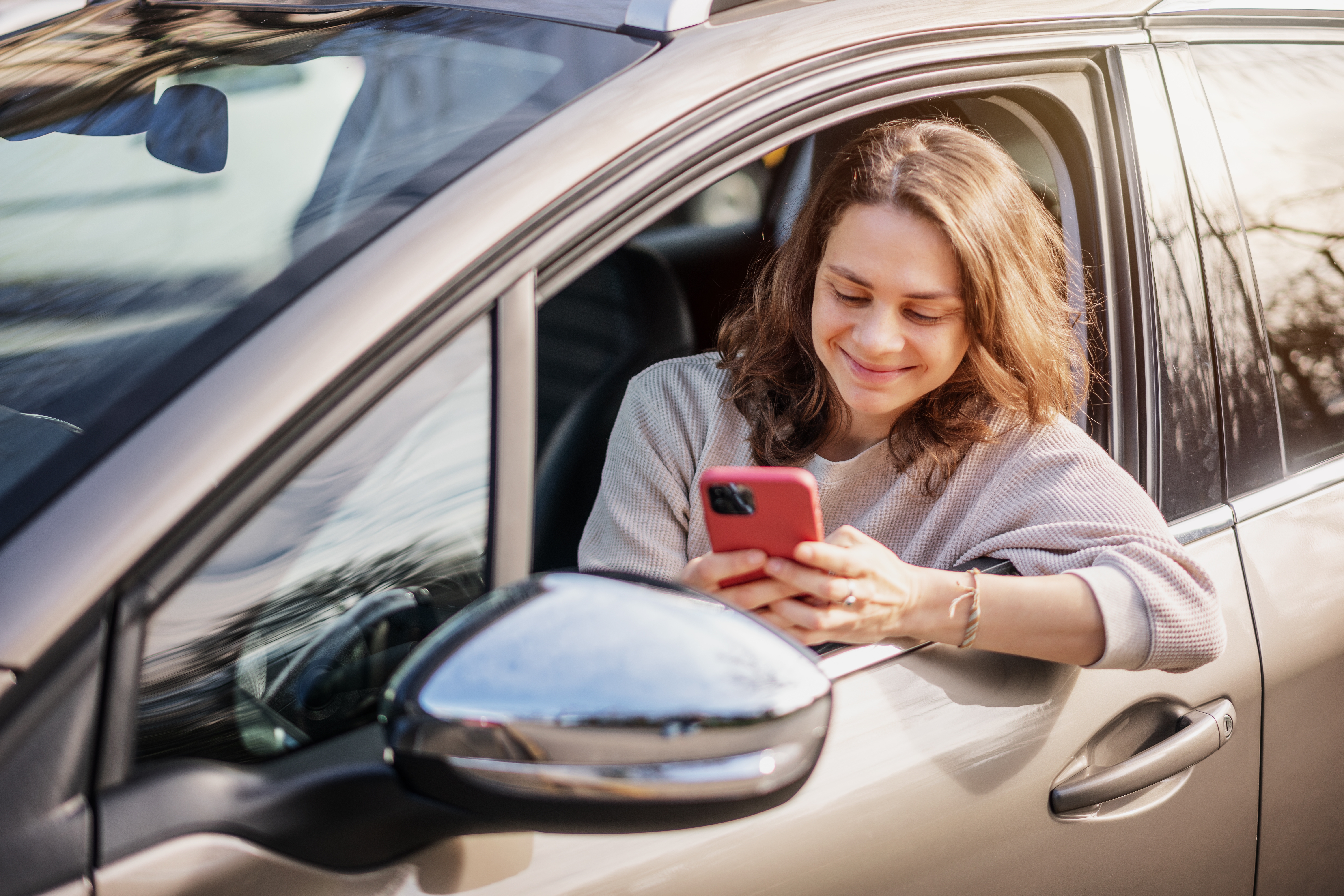 Cheerful young woman sitting in a car in the driver's seat looking into a smartphone, paying for parking and navigating in the city | Source: Shutterstock