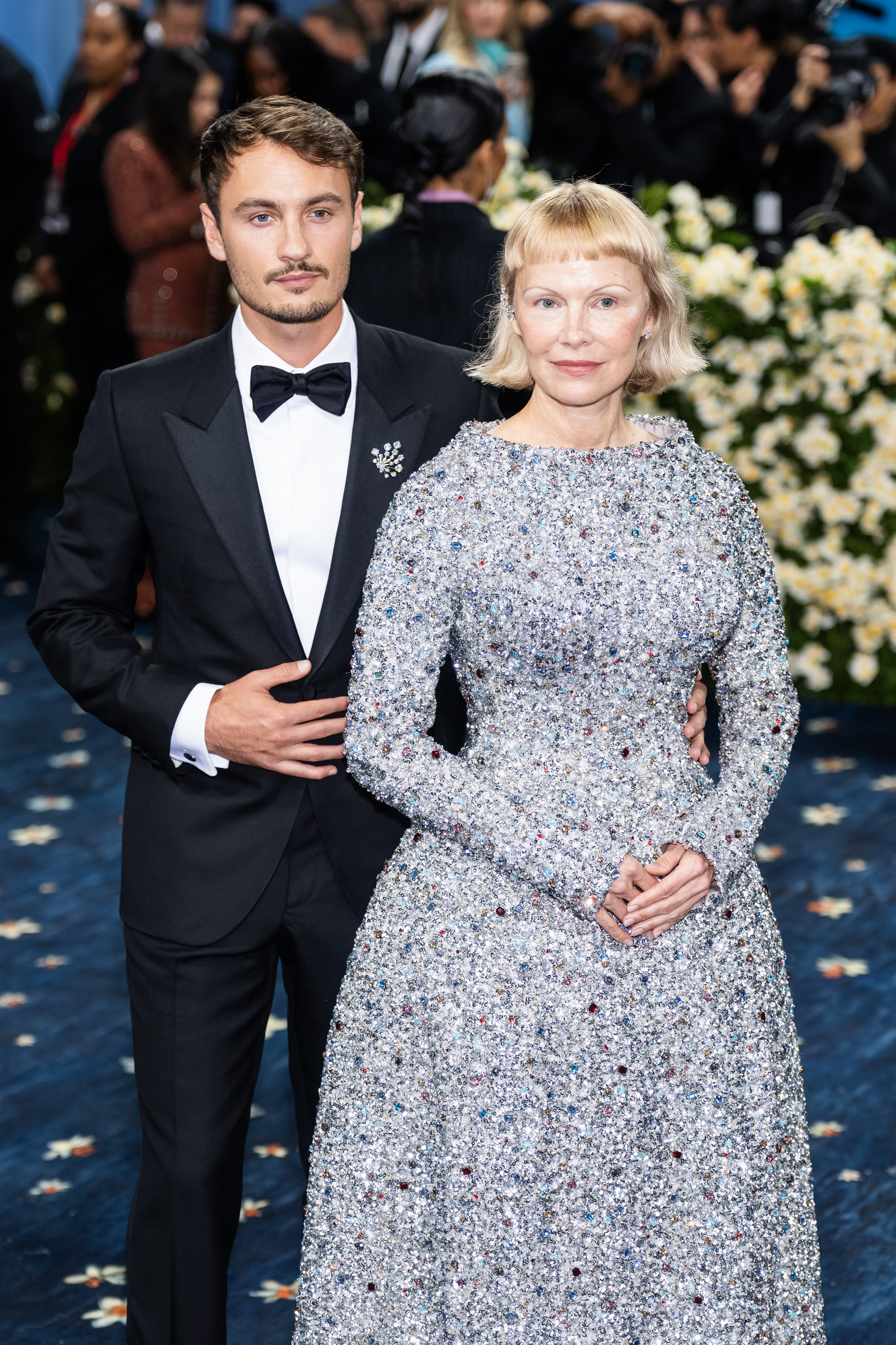 Pamela Anderson and Brandon Thomas Lee share a composed moment on the blue carpet at the 2025 Met Gala. Anderson stands confidently in a dazzling gem-covered gown, while Lee gently rests his hand on her back, both maintaining calm and poised expressions amid a flurry of flashing cameras.
