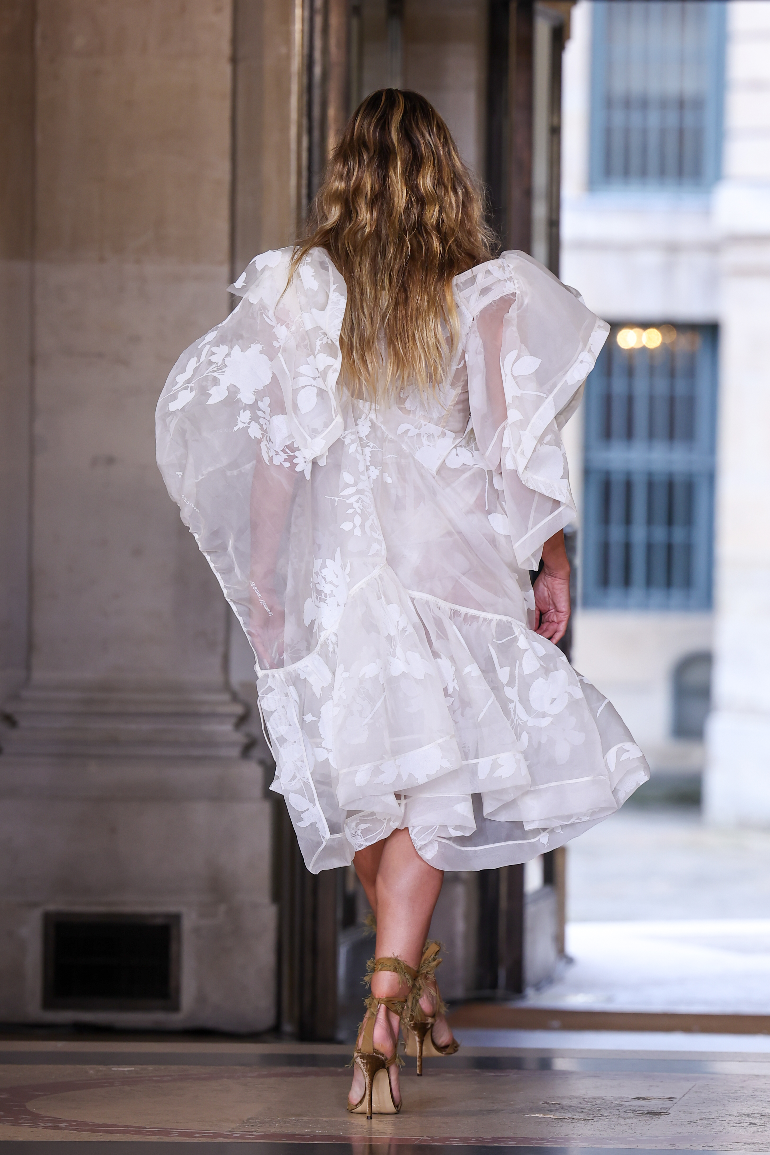 A feather-inspired, rhinestone-encrusted gauze detail cascaded down the front of Heidi Klum’s legs, completing the dazzling design. She paired the look with tan sandal heels featuring frayed strings wrapped around her ankles, enhancing the outfit’s artfully undone effect.
