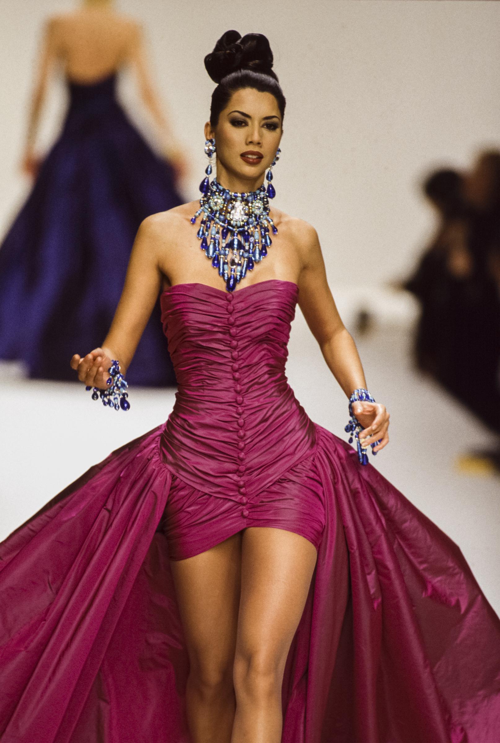 A model showcases the original Jean-Louis Scherrer design on the runway in 1994, wearing the fuchsia strapless gown with ruching and a dramatic train. The look is styled with bold statement jewelry, including a large jeweled necklace, earrings, and bracelets, highlighting the couture detail of the vintage piece later worn by Amal Clooney.