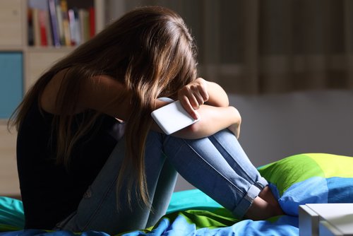 Niña sentada sobre su cama con gesto triste y el celular en su mano. | Foto: Shutterstock