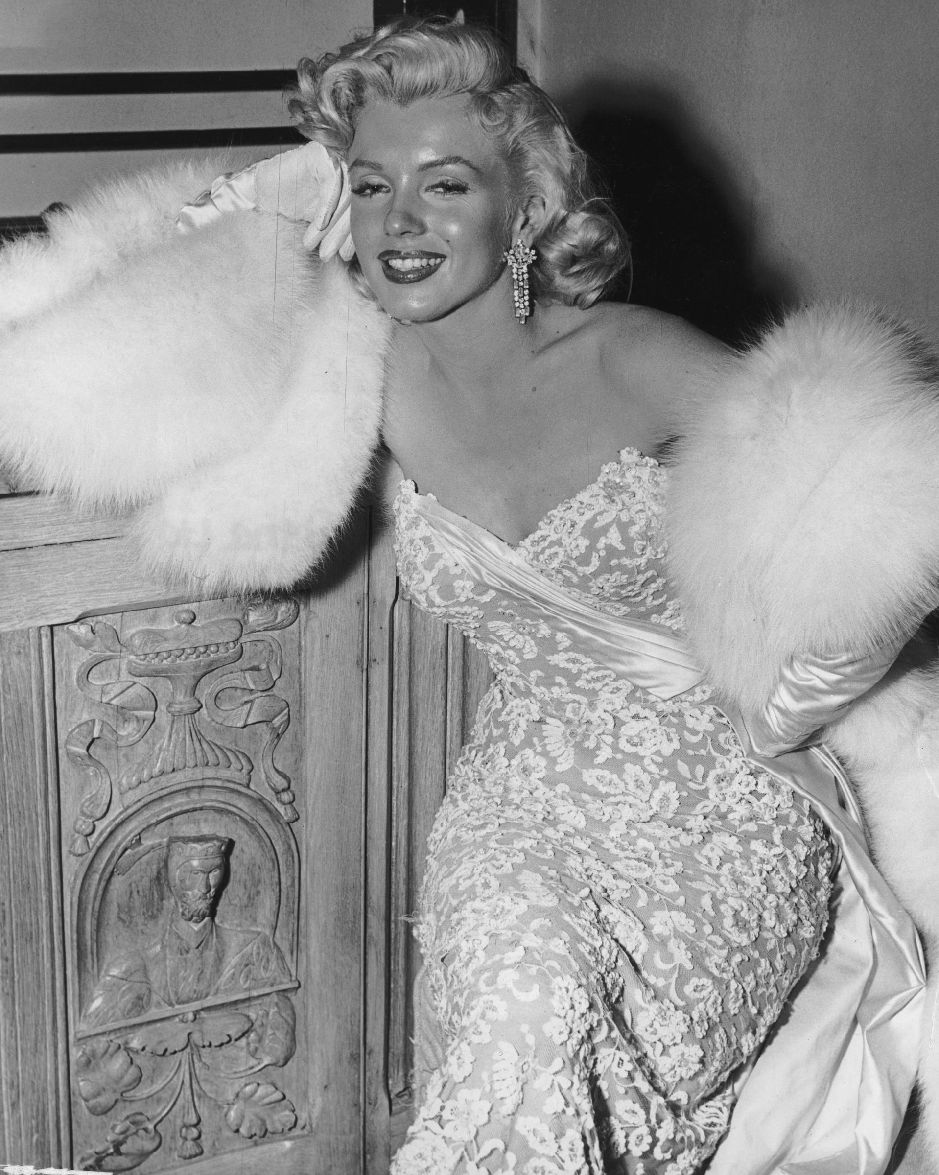 Marilyn Monroe (1926 - 1962), wearing white fur wrap and strapless evening gown, leans on a wooden cabinet during a party for the film, "How To Marry A Millionaire" at director Jean Negulescu's house, circa 1953, in Hollywood, California. | Source: Getty Images
