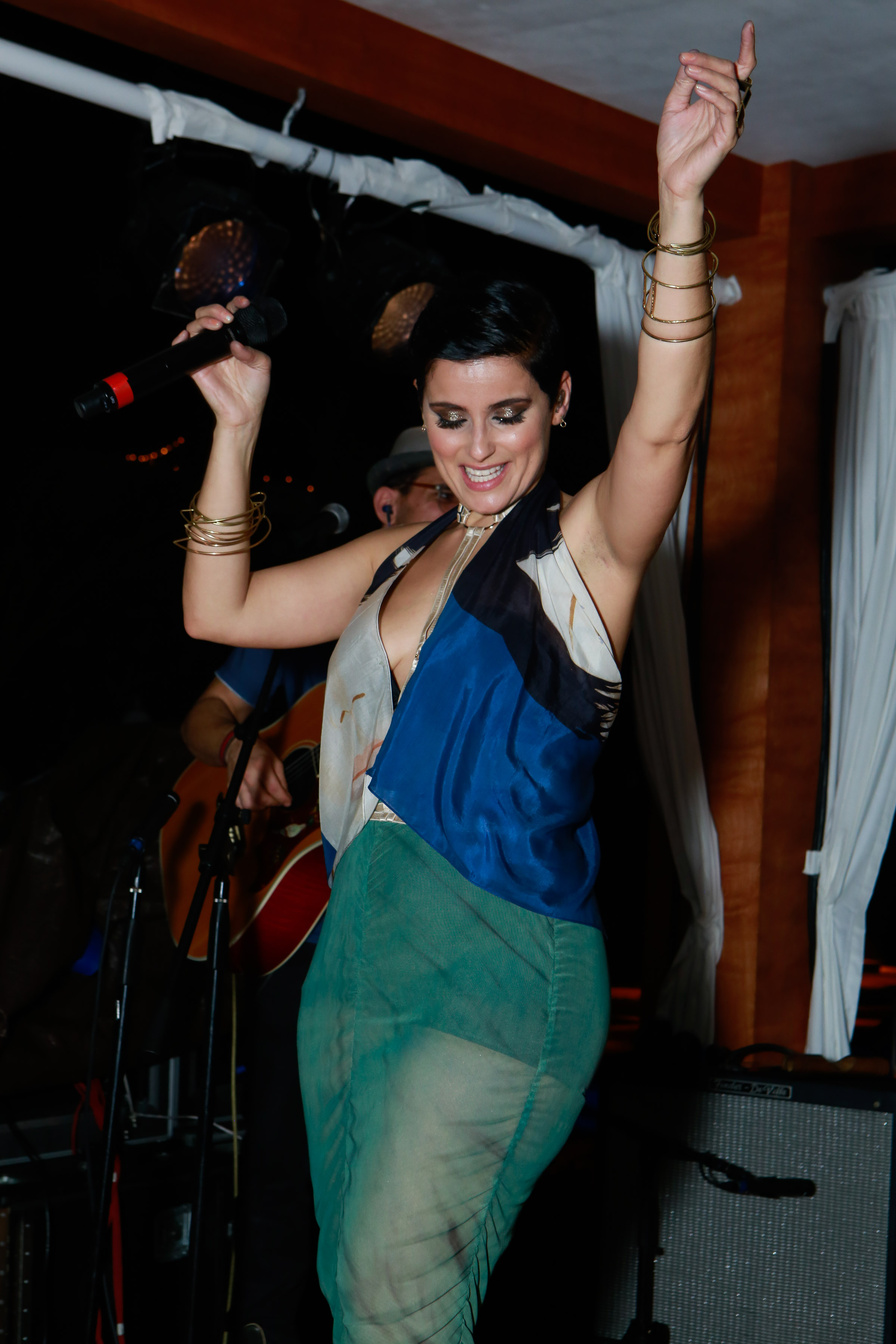 Furtado performs on stage wearing a multicolored halter-style top and sheer green trousers. Her hair is styled short, and she accessorizes with stacked gold bangles and metallic eye makeup.